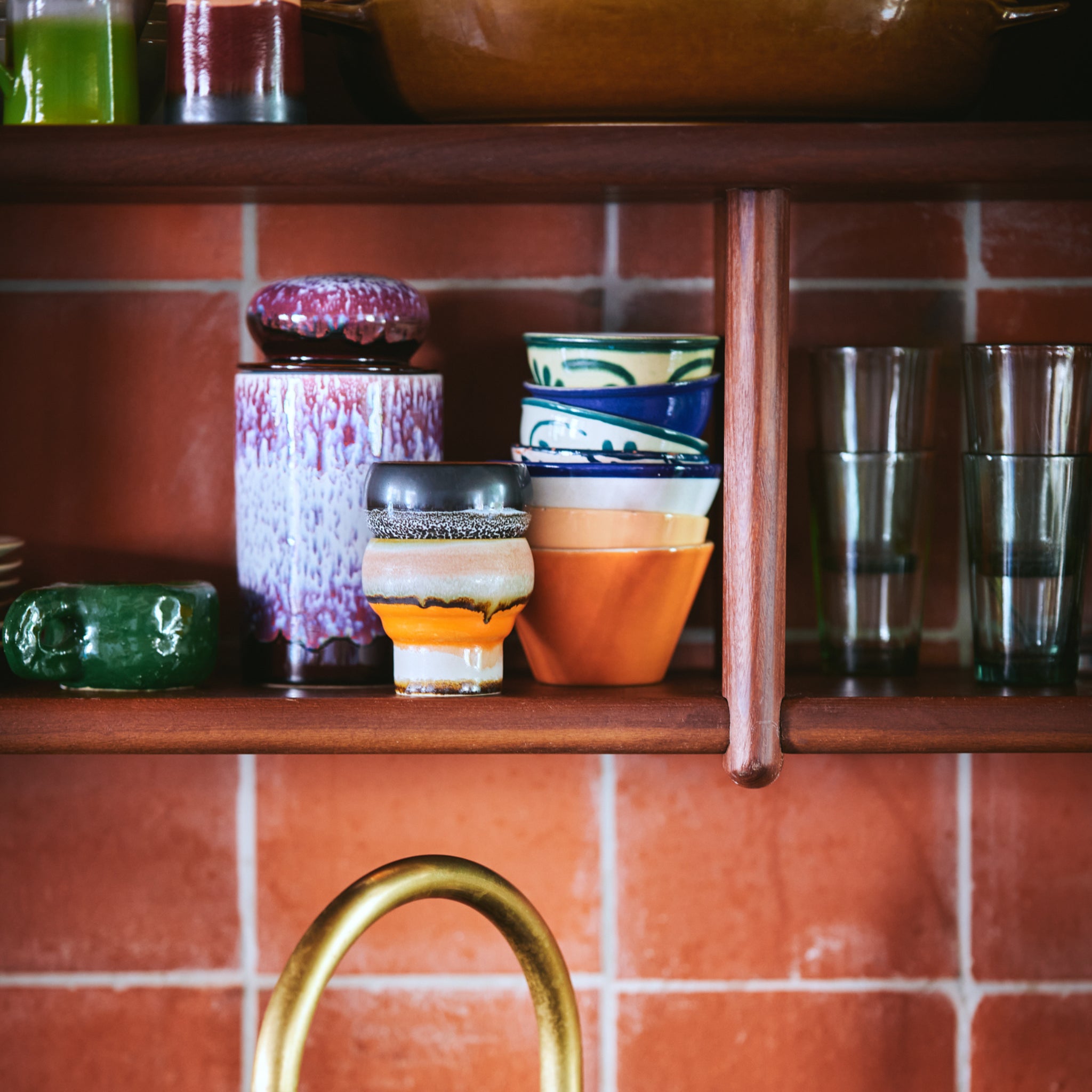 Op een houten keukenplank tegen een roodbruine betegelde muur staan kleurrijke kopjes, kommen, een groene schaal, heldere glazen en een HK Living 70s Ceramics Voorraadpot Yeti; onderaan staat een koperen kraan.