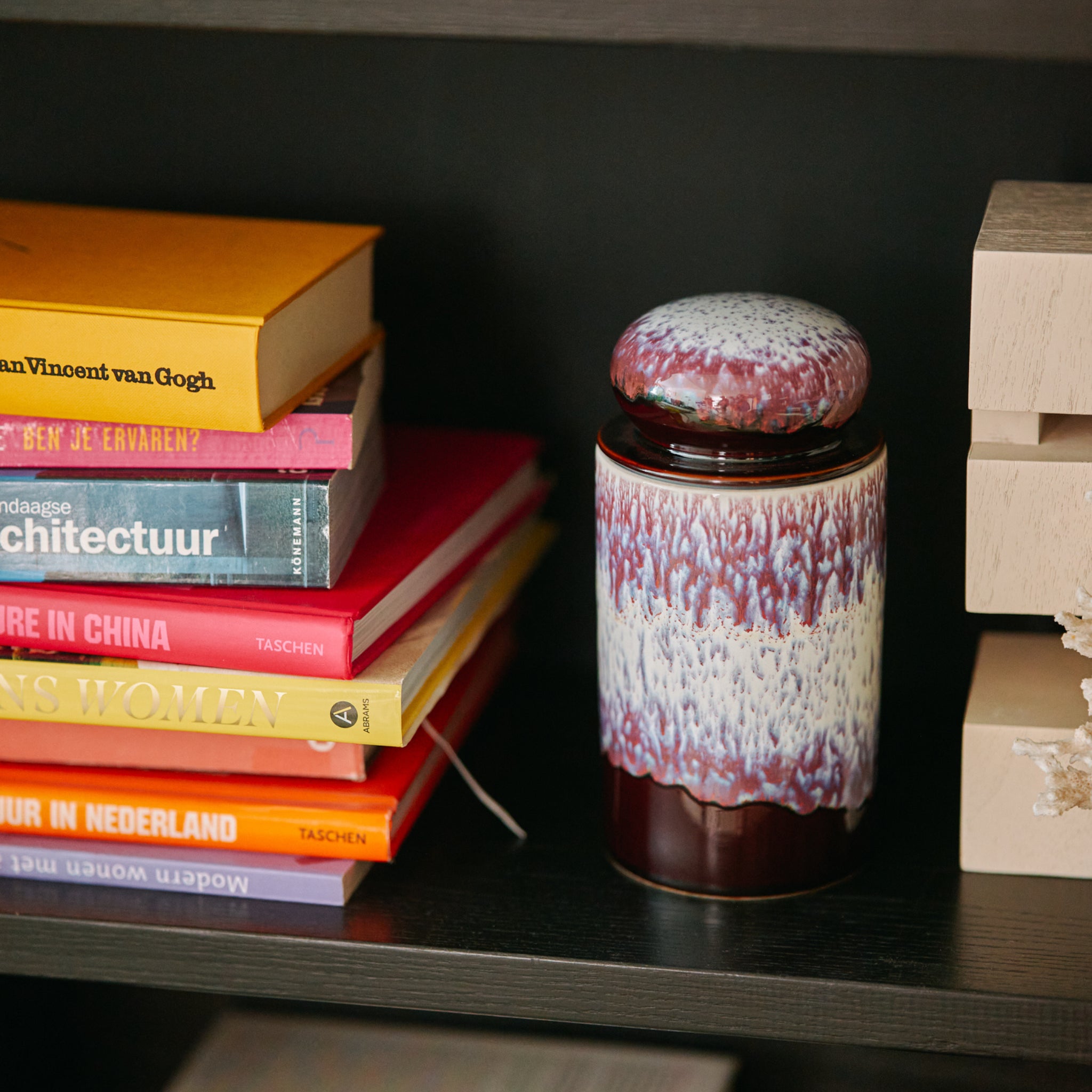 De HK Living 70s Ceramics Voorraadpot Yeti, met een gestructureerd kastanjebruin en wit deksel, staat op een donkere plank naast kleurrijke boeken, waaronder een geel Van Gogh boek en verschillende architectuur- en reistitels.