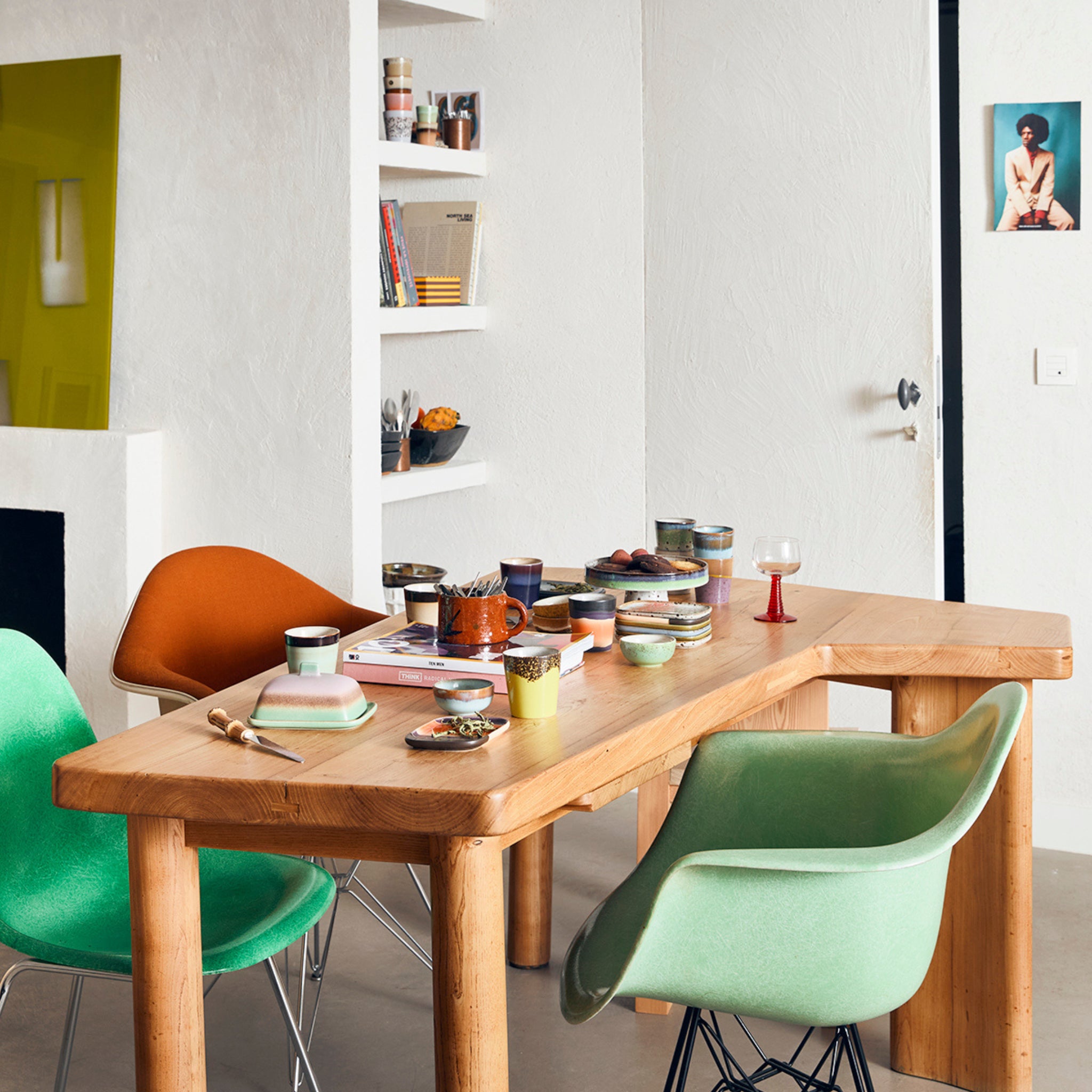 Een moderne eethoek met een houten tafel, kleurrijke stoelen en gezellige inrichting met HK Living 70s Ceramics XS Kom Castor 4 st. schalen op tafel, planken met boeken en decor, en een foto bij een open deur.