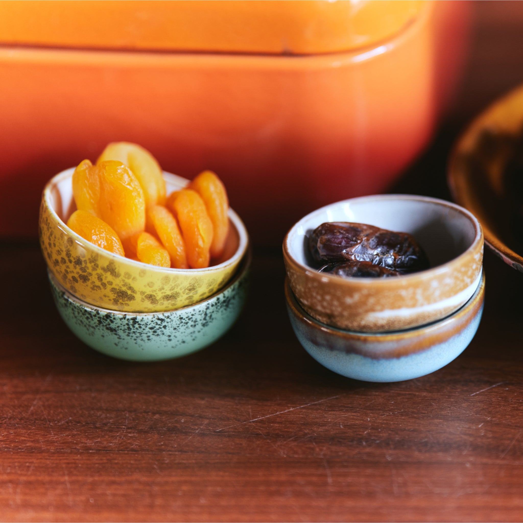 Twee kleine schaaltjes uit de HK Living 70s Ceramics XS Kom Castor set rusten op een houten oppervlak; in de ene zitten gedroogde abrikozen, in de andere dadels. Een oranje HK Living schaaltje zorgt voor een vleugje jaren 70 keramiek op de achtergrond.