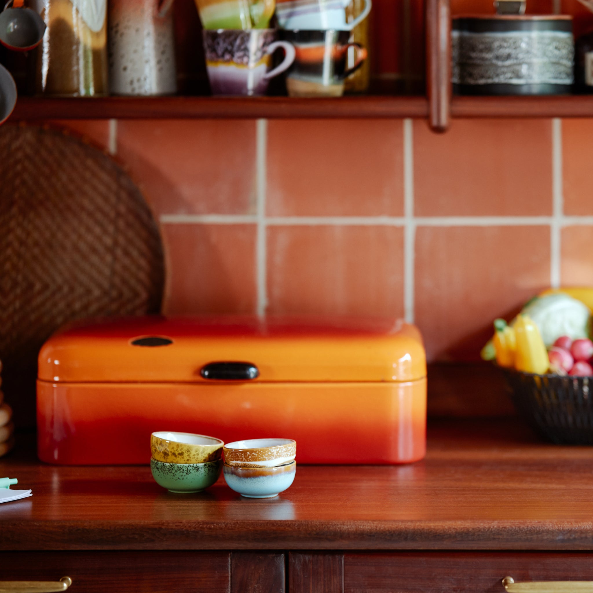 Twee kleine HK Living 70s Ceramics XS Kom Castor kopjes staan op een houten aanrecht voor een oranje brooddoos, met fruit en gestapelde kopjes op planken tegen een betegelde keukenmuur op de achtergrond.