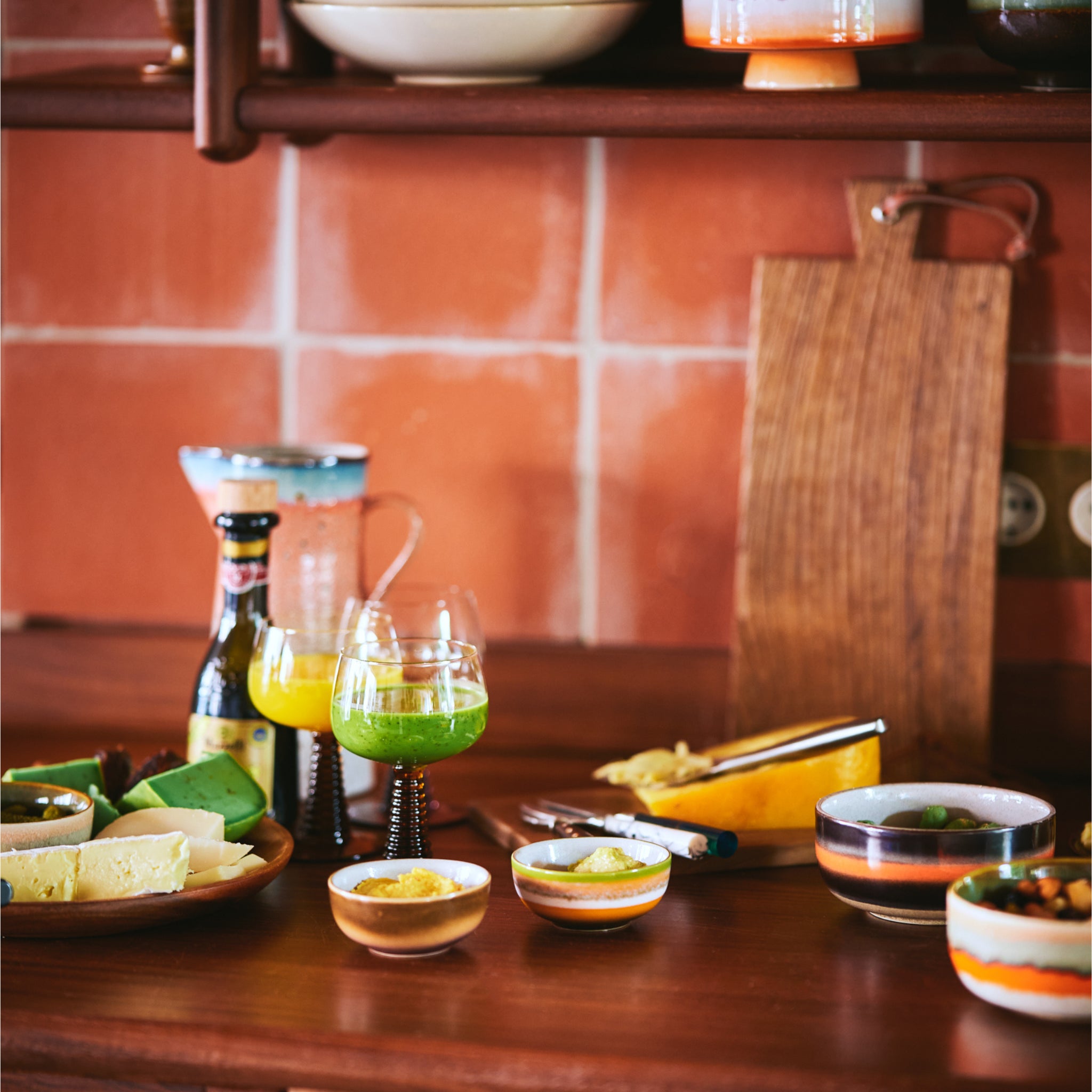Een houten aanrecht toont HK Living 70s Ceramics XS Kom Sierra 4 st., elk gevuld met kaas, groene saus en gele dip. Twee kleurrijke drankjes, een bierfles, kruik, keukengerei en snijplank staan tegen een betegelde muur.