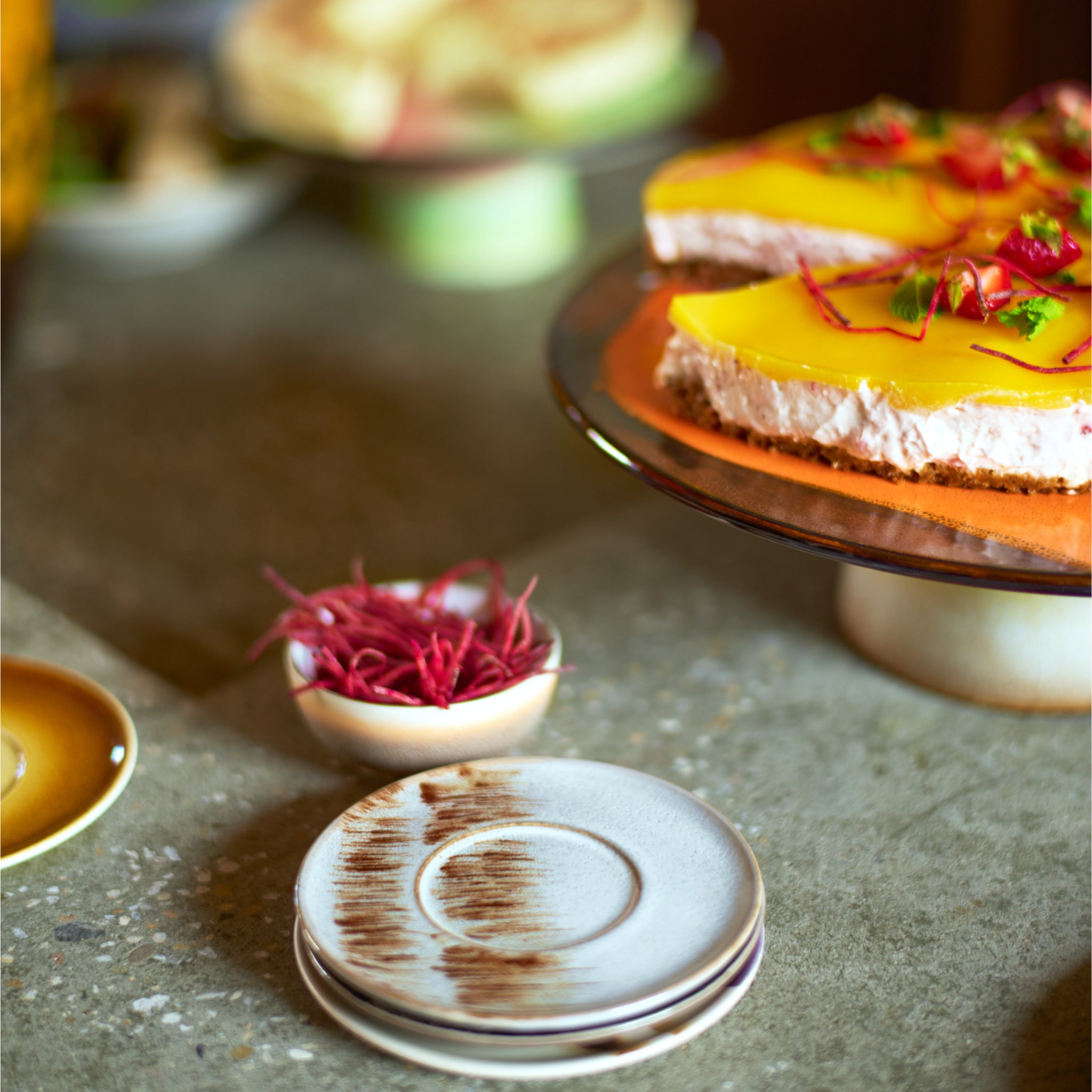 Een set HK Living 70s Ceramics XS Kom Sierra 4 st. borden rust op een stenen oppervlak naast een schaal met rode garnering, met op de achtergrond een geelgeglazuurde, met kruiden bedekte cake op een standaard.