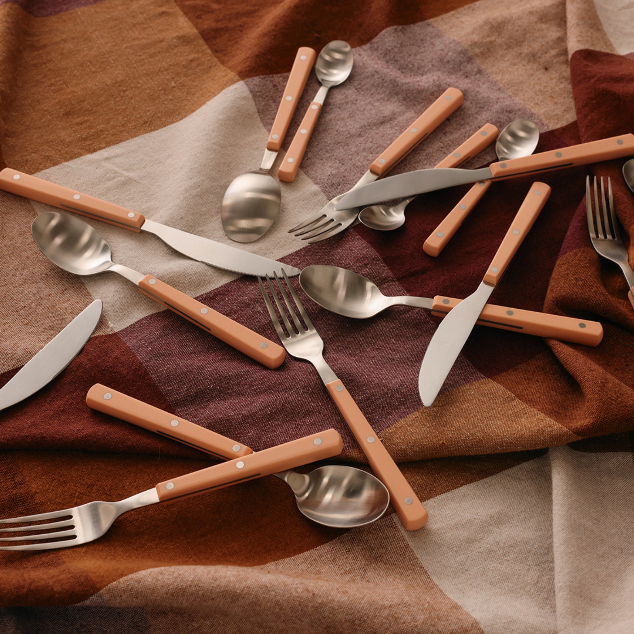 De HK Living 70s Tafelmes Perzik heeft een lichtbruin handvat, hier afgebeeld naast verschillende vorken en lepels op een geruite bruine, beige en kastanjebruine stof.