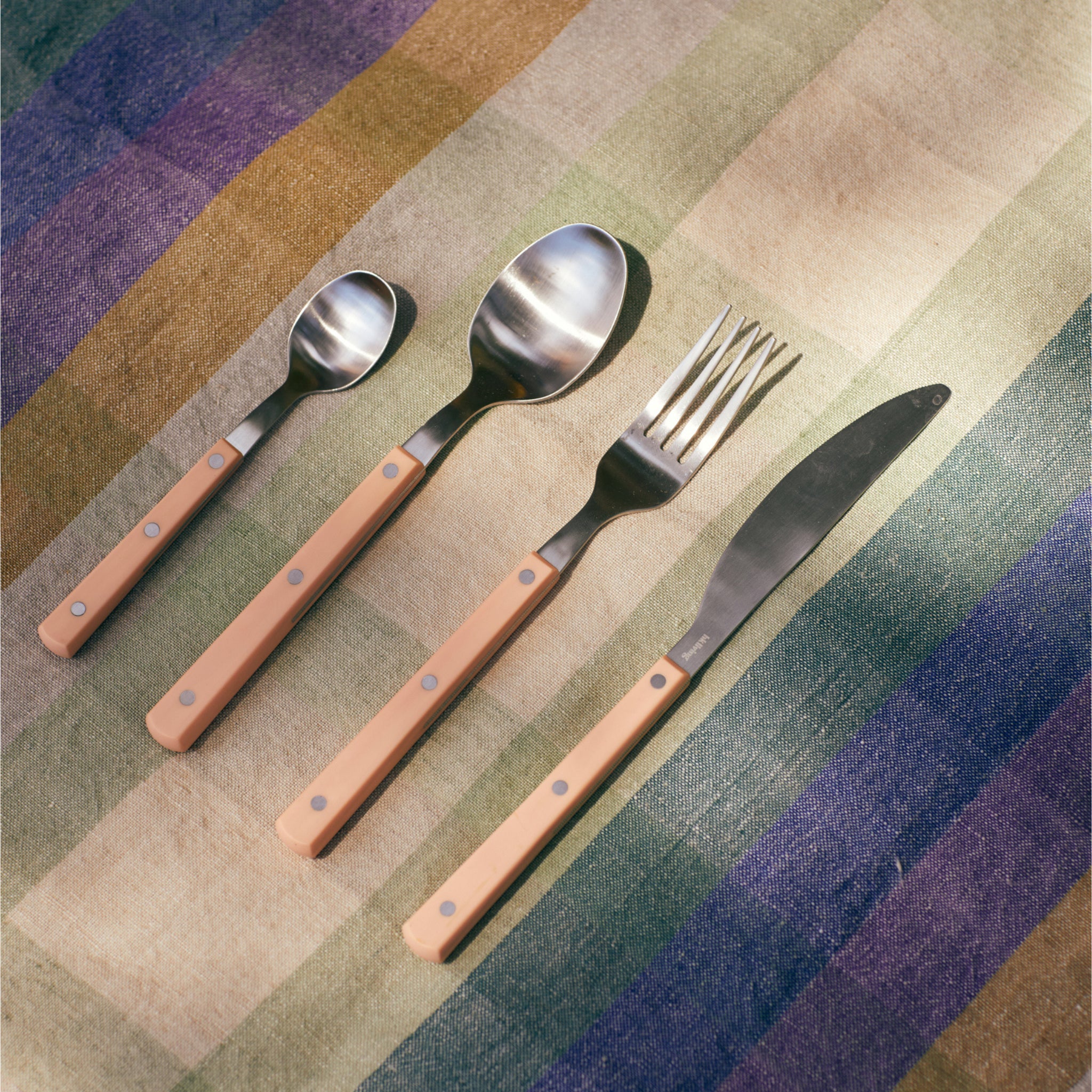 Een HK Living 70s Cutlery tafelvork perzik met een lichtbruin handvat ligt netjes op een gestreept, veelkleurig tafelkleed in het zonlicht.