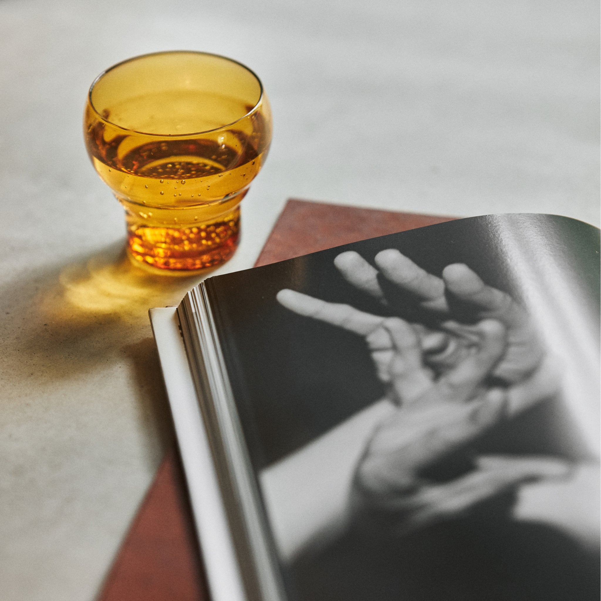 De HK Living 70s Glassware Bulb Glazen Amber (set van 4) staat op een licht oppervlak naast een open boek met een zwart-wit foto en roept vintage charme op met zijn amberkleurige glas en retro stijl.