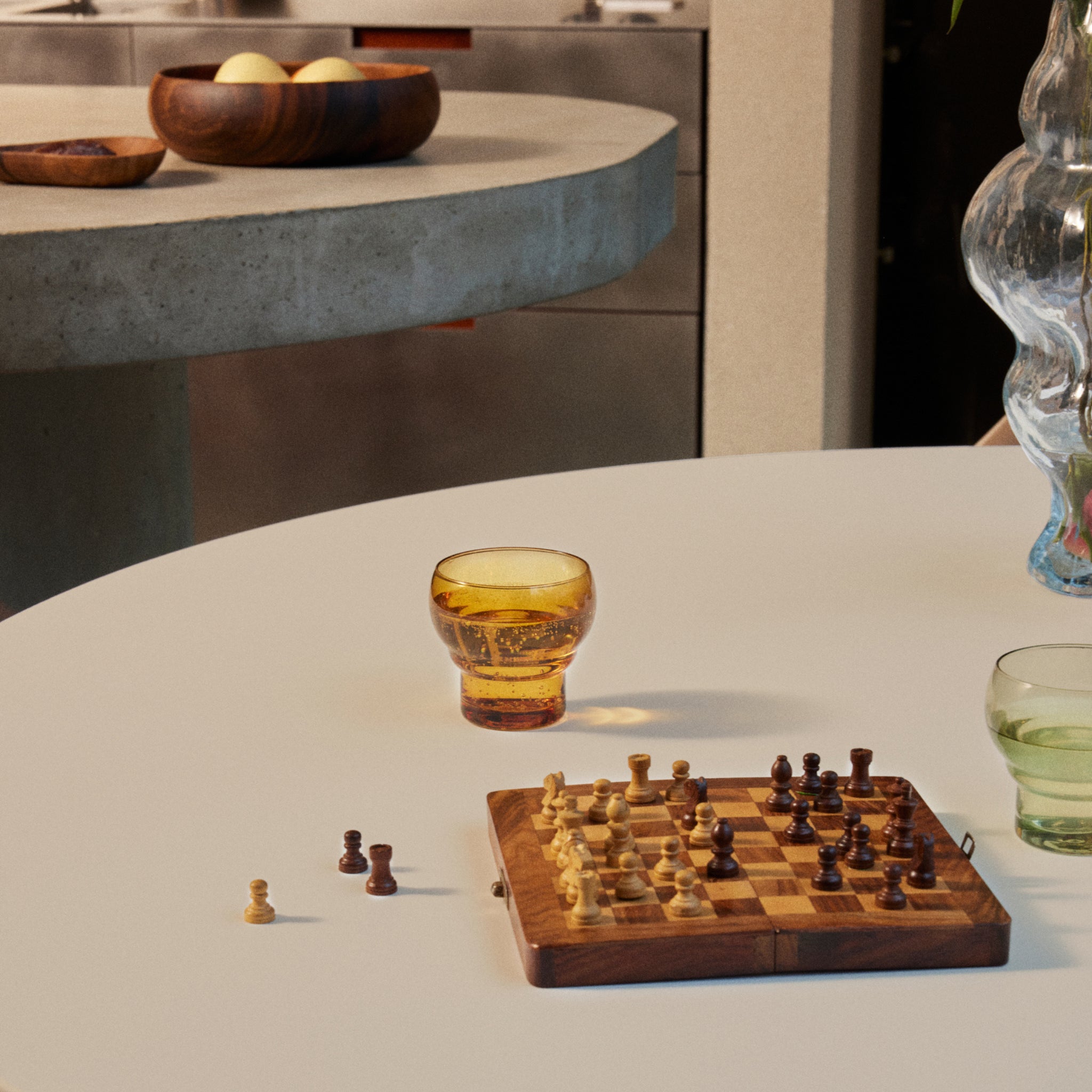 Een klein schaakbord met stukken staat op een ronde witte tafel naast twee HK Living 70s Bulb Glazen Amber glazen - een met een gouden glaasje, een groenig. Een houten fruitschaal staat op het betonnen aanrecht op de achtergrond.