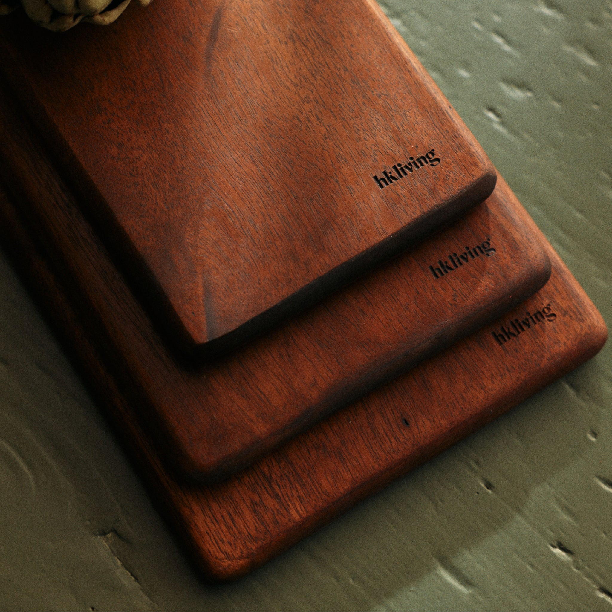Drie HK Living Acacia Snijplanken 60/15 van verschillende afmetingen zijn gestapeld op een groen gestructureerd oppervlak. In elke hoek is het hkliving logo gegraveerd, wat een stijlvolle touch geeft aan je keukenbenodigdheden.