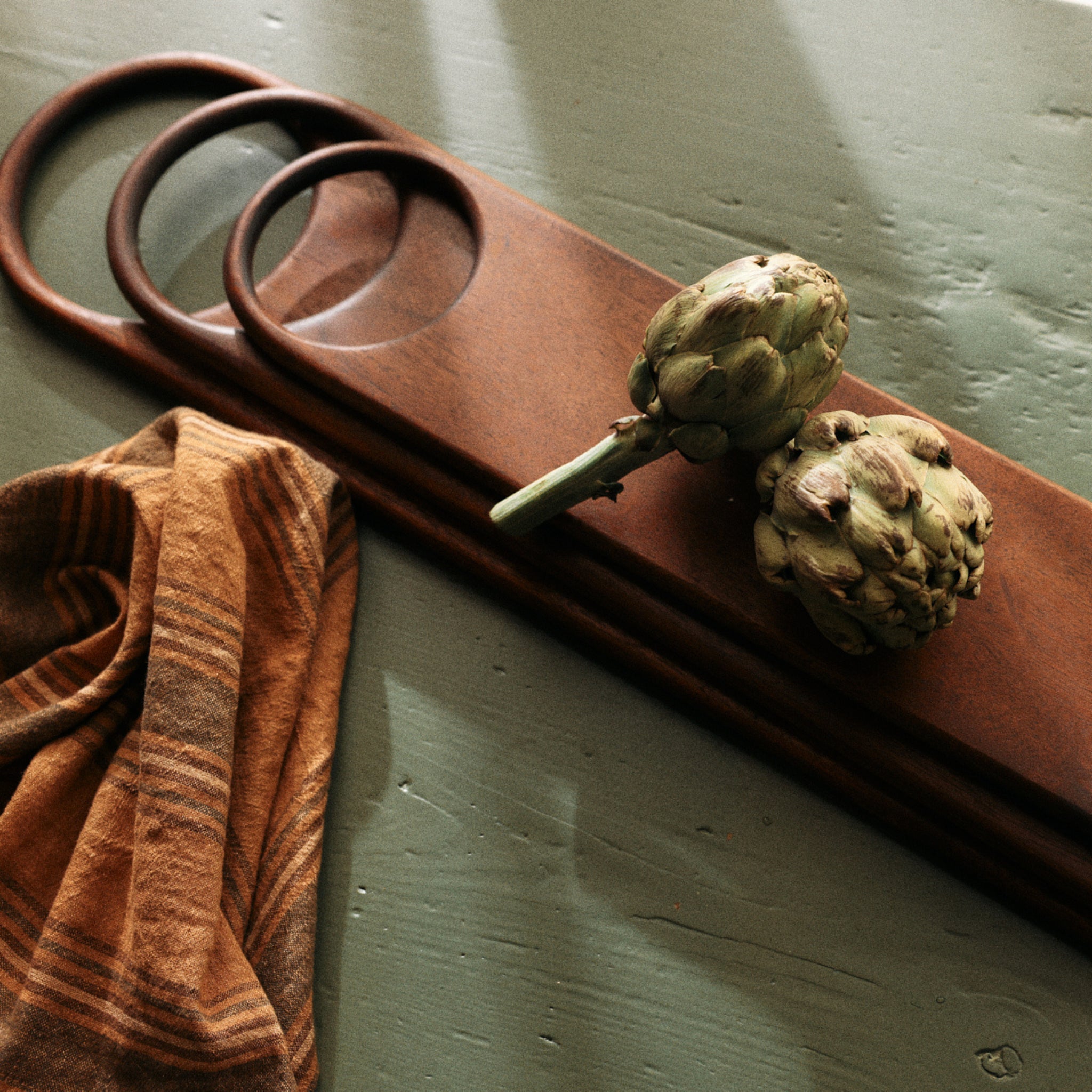 Twee artisjokken zitten op de HK Living Acacia Snijplank 60/15. Een gestreepte bruine doek rust ernaast op een groen, versleten oppervlak terwijl zacht licht zachte schaduwen werpt over het acaciahout.