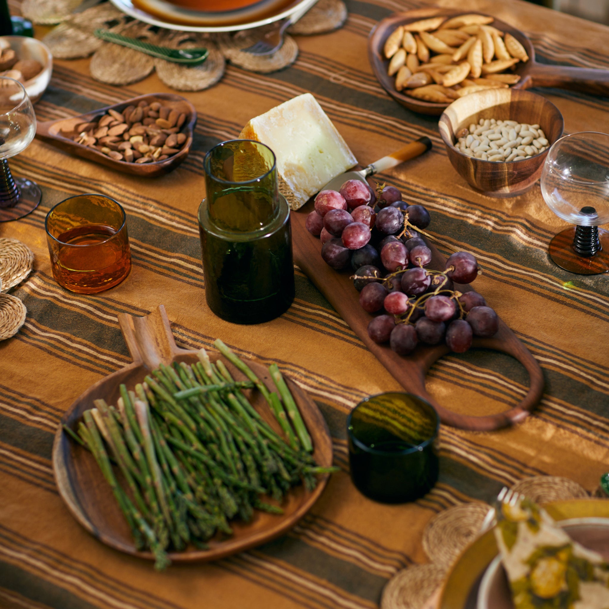 Een houten tafel gedekt met borden met asperges, druiven, kaas, amandelen, broodstengels, pasta en kleurrijke drankjes staat naast de HK Living Acacia Snijplank 60/15 van HK Living en creëert een warme en uitnodigende eetsfeer.