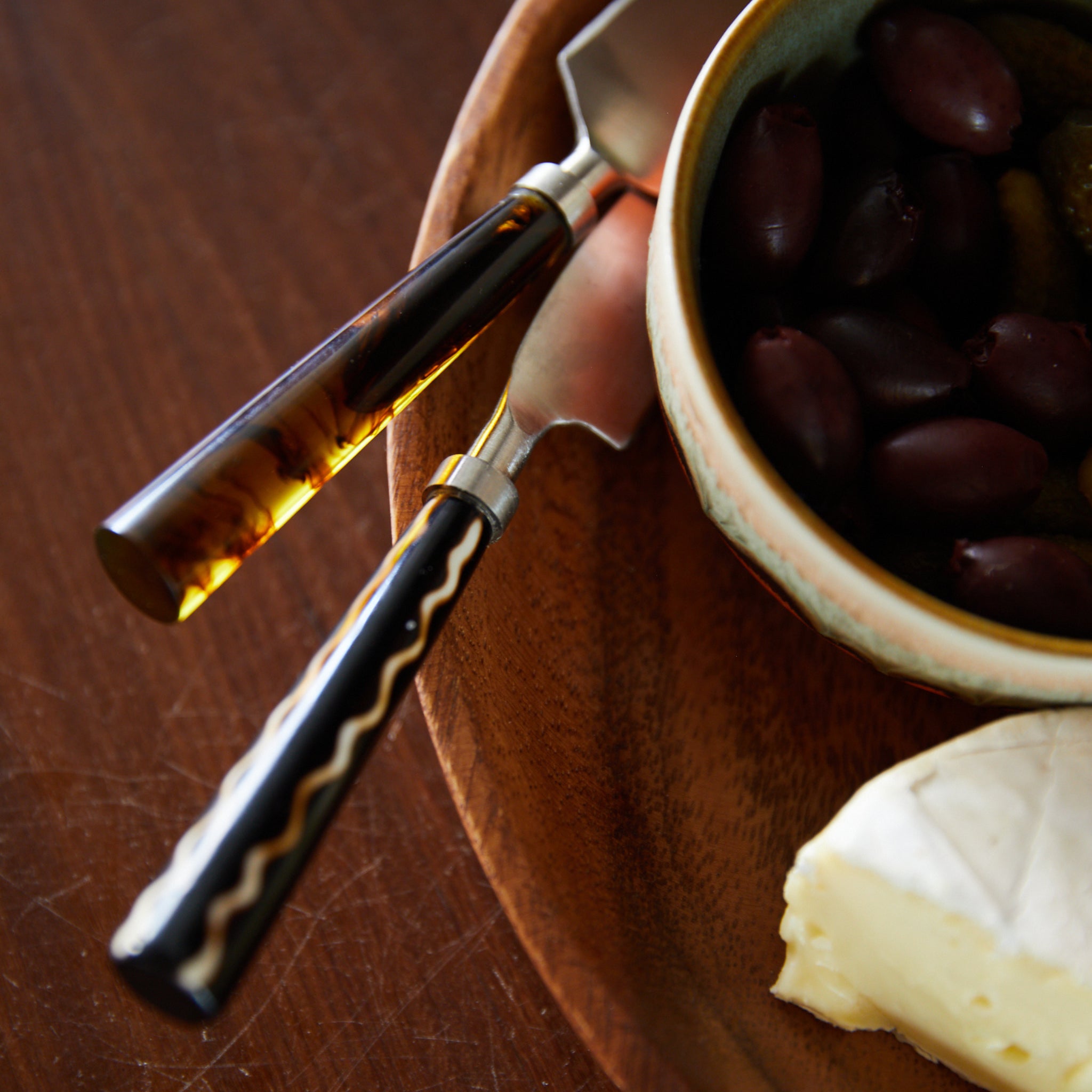 Een close-up van een houten schaal met daarop HK Living Kaasmesjes Havana 3 st., een vork, een schaaltje zwarte olijven en een plakje zachte kaas, kunstig gerangschikt op een donkere houten tafel.