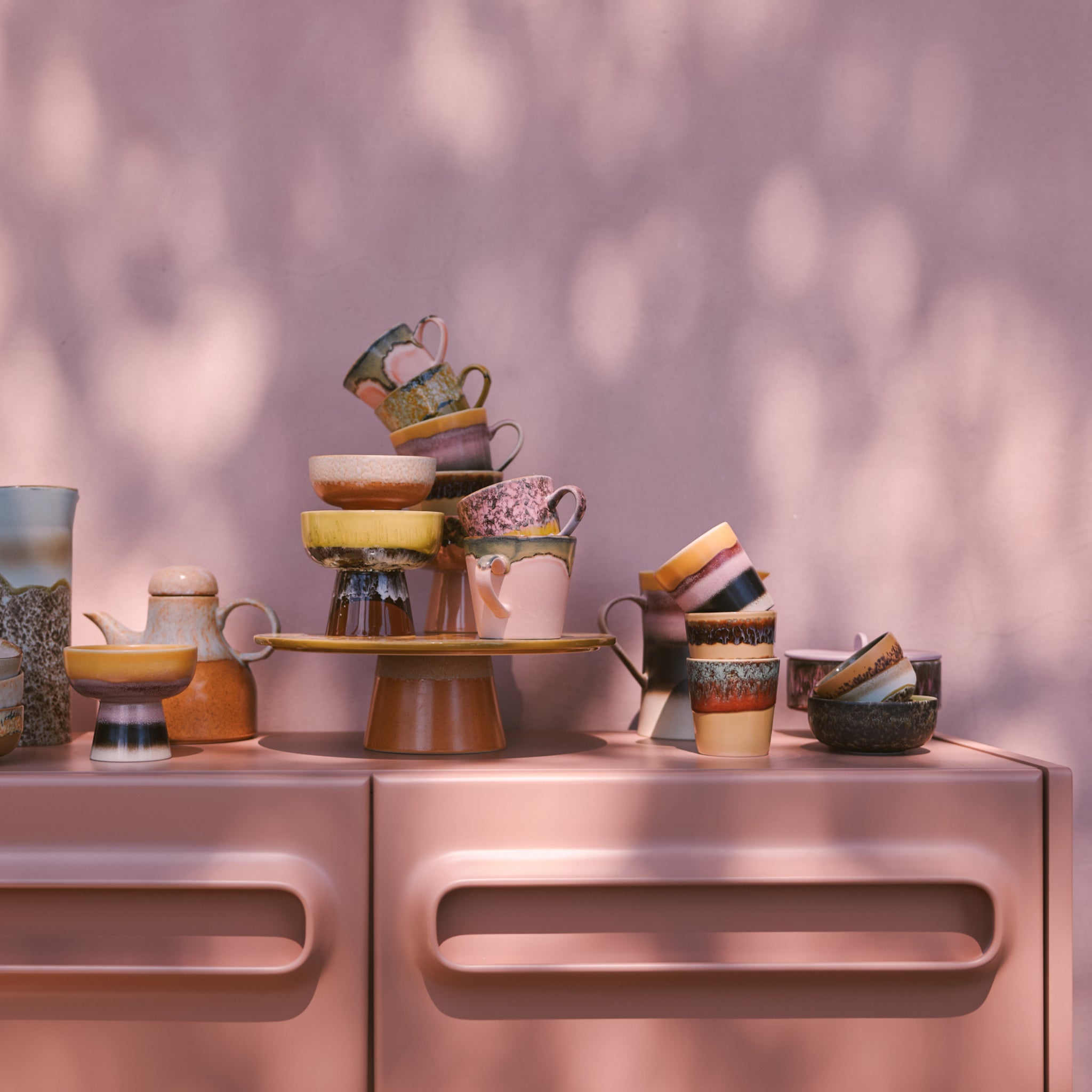 Een stapel HK Living 70s Ceramics Koffiekop Cliffs kopjes staat op een roze kast tegen een roze muur, met zacht gedimd zonlicht dat retro stijl schaduwen toevoegt aan het kleurrijke tafereel.