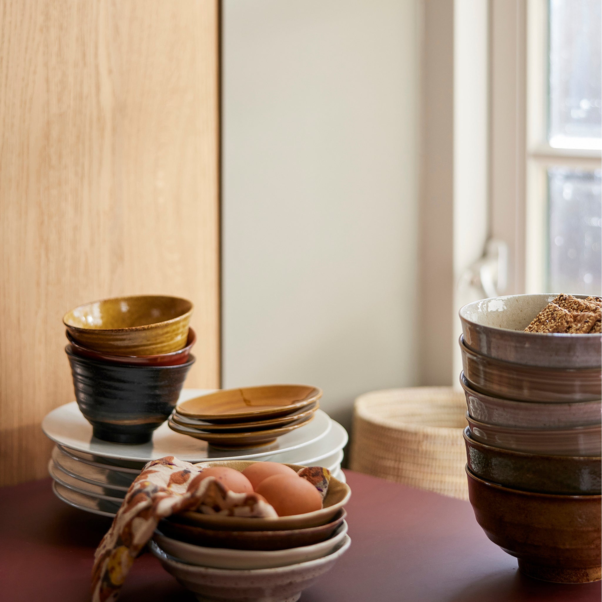 Een stapel HK Living Kyoto Ceramics Japans Bord D14 kommen en borden staat op een tafel bij het raam, met een doek en drie bruine eieren ernaast. Zacht natuurlijk licht schijnt door het raam.
