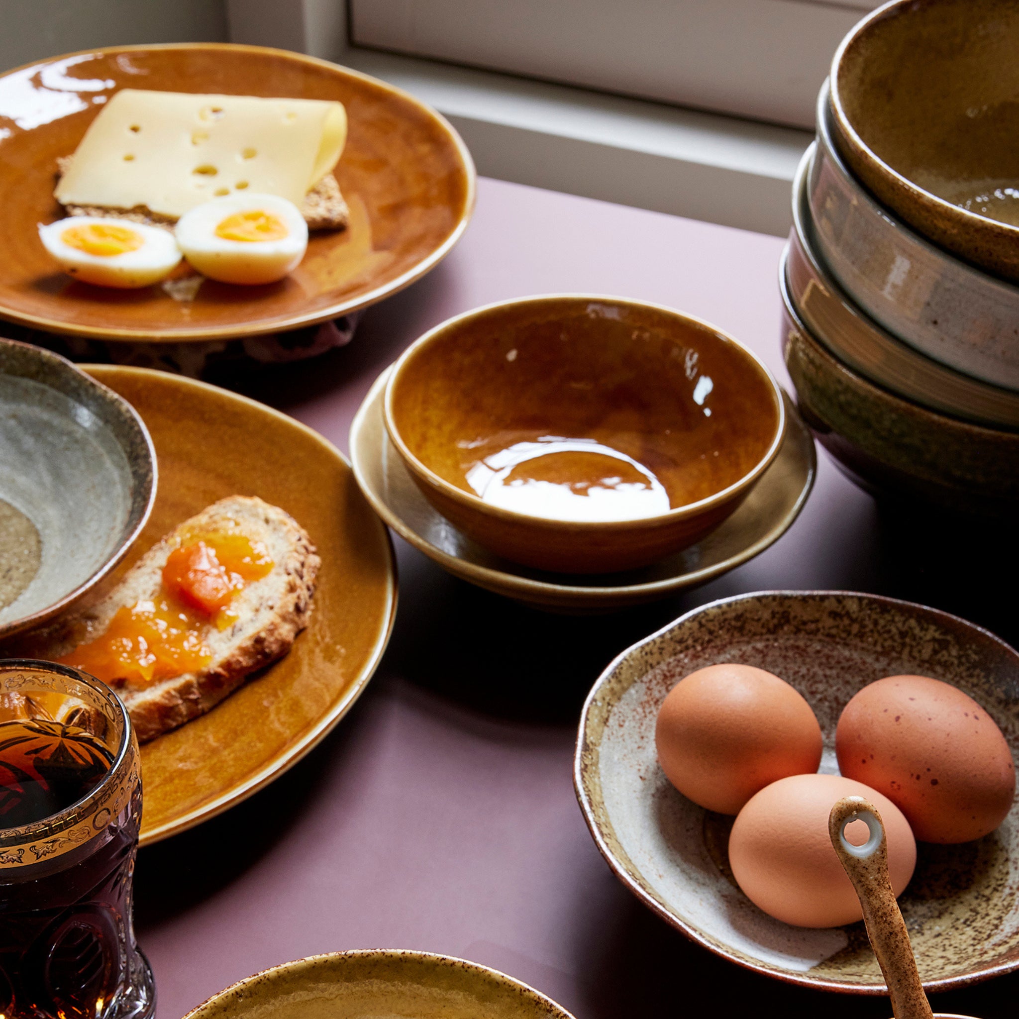 Een gedekte ontbijttafel met kommen, kaas, gekookte eieren, toast met sinaasappeljam, bruine eieren en een donker drankje op de HK Living Kyoto Ceramics Japans Bord D25 van HK Living op een mauve ondergrond bij een raam.