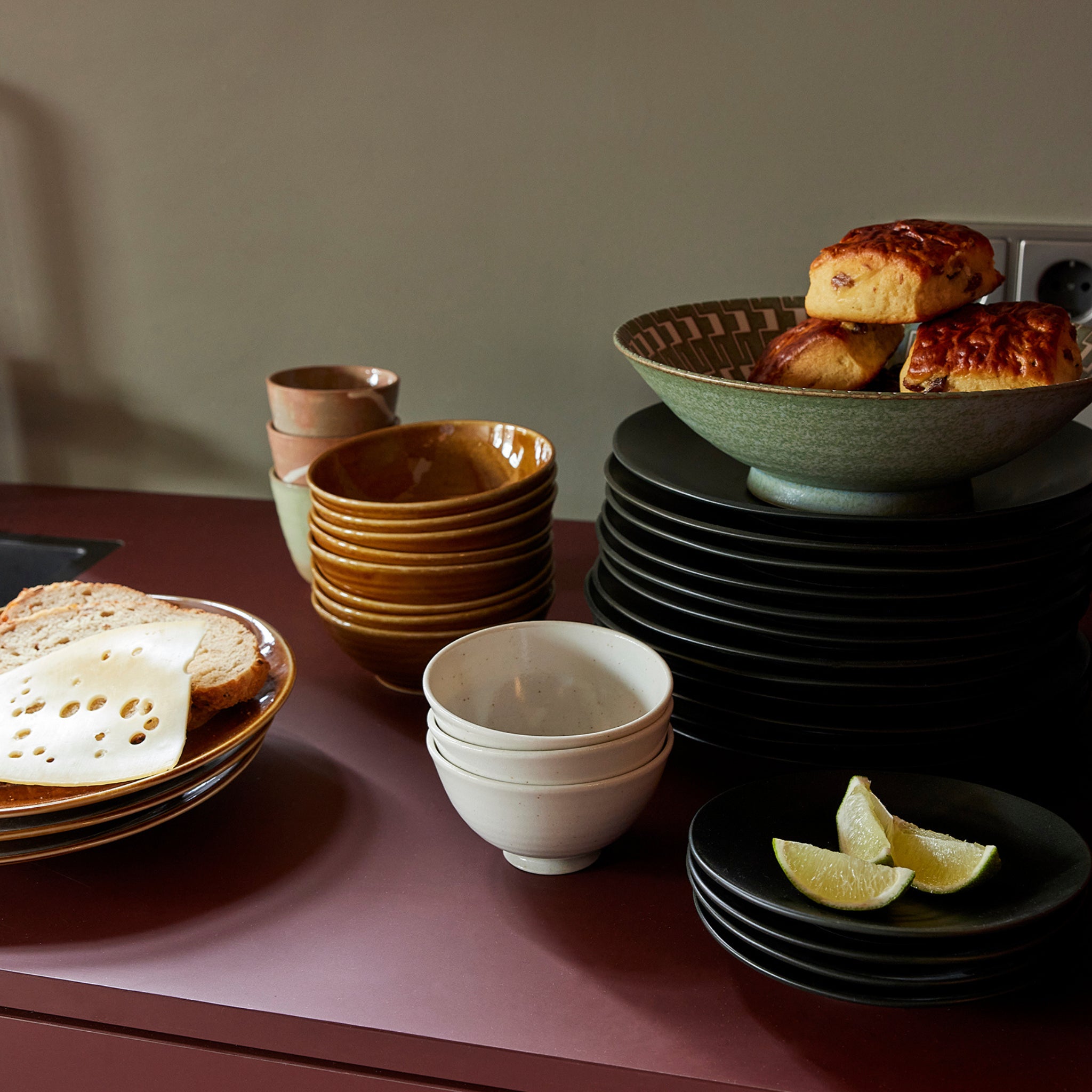 Op een aanrecht staan HK Living Kyoto Ceramics Japanse Rijstkom Wit Gespikkeld schalen gevuld met brood, gebak en citroenpartjes. Stapels porseleinen borden en kopjes staan op een roodbruin oppervlak.