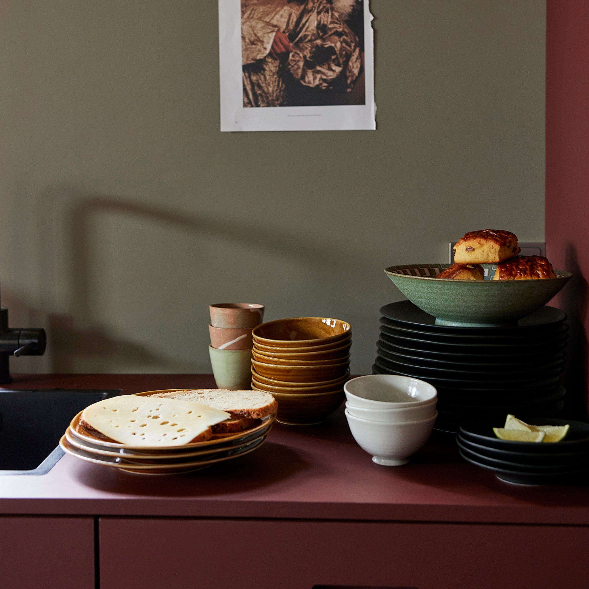 Op een mauve aanrecht staan HK Living Kyoto Ceramics Japanse Rijstkom Wit Gespikkeld kommen gestapeld met borden platbrood, kopjes, gebak in een kom en partjes limoen; op de taupekleurige muur erachter is een zwart-witfoto afgeplakt.