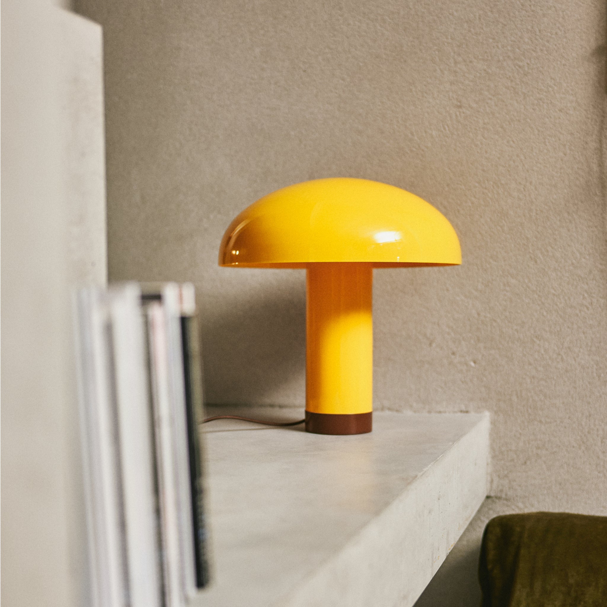 Een HK Living Lounge Tafellamp Butterscotch met een gele paddenstoelkap en bruine voet staat op een betonnen plank naast rechtopstaande boeken tegen een beige muur. De lamp is compatibel met dimbare E27 LED-lampen voor aanpasbare verlichting.