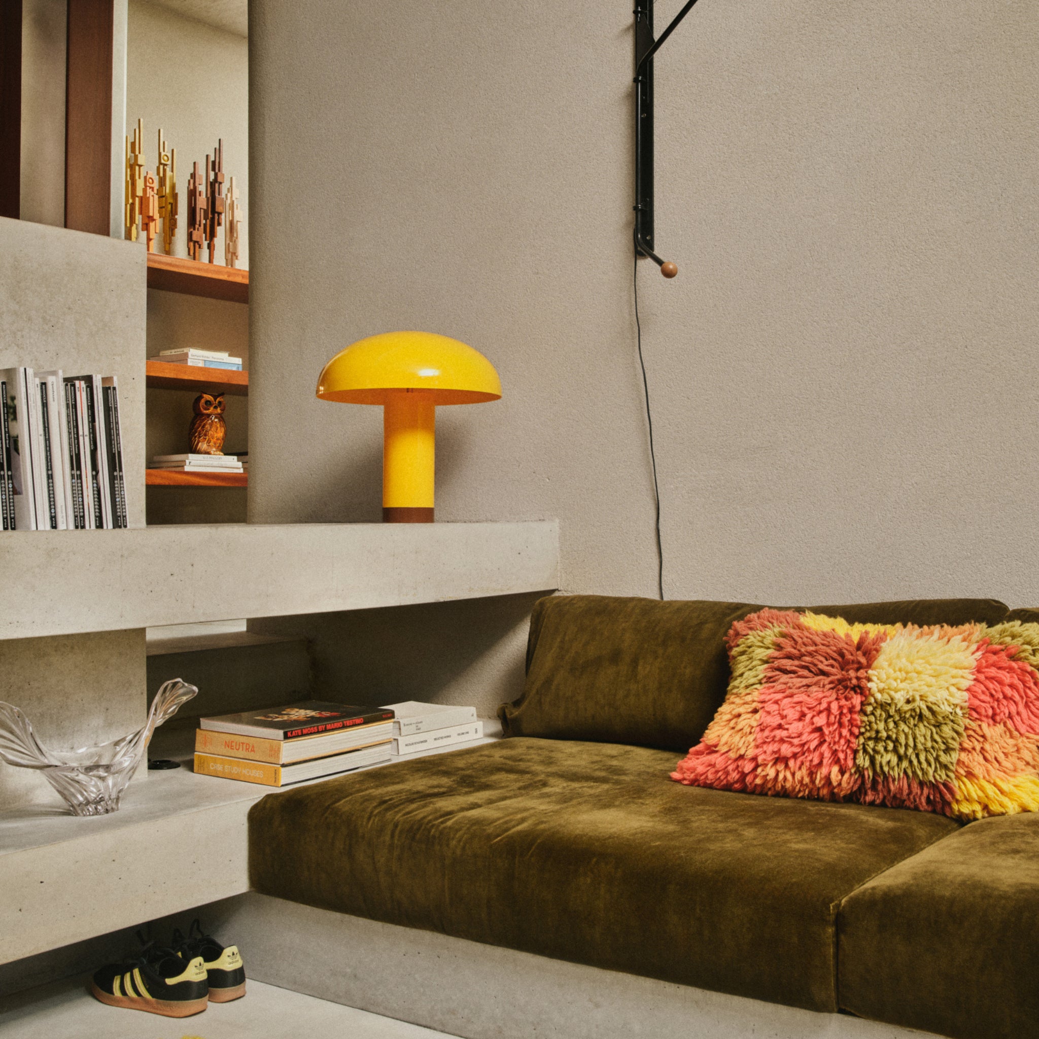 Een gezellige moderne woonkamer met een groen fluwelen bank, kleurrijk shaggy kussen en een HK Living Lounge Tafellamp Butterscotch van HK Living met een dimbare E27 LED op een betonnen plank, omringd door boeken, decorstukken en sneakers op de vloer.