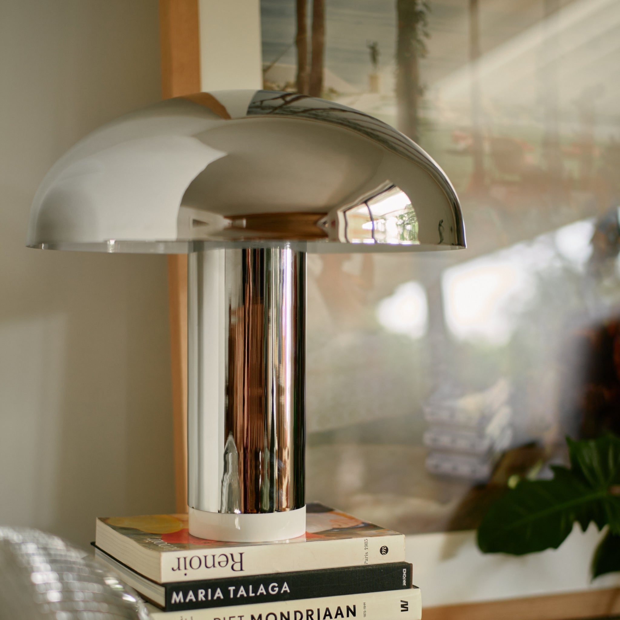 De HK Living Lounge Tafellamp Chroom, een moderne koepelvormige lamp van HK Living, ligt op een stapel boeken voor ingelijste kunstwerken en een groene bladplant.