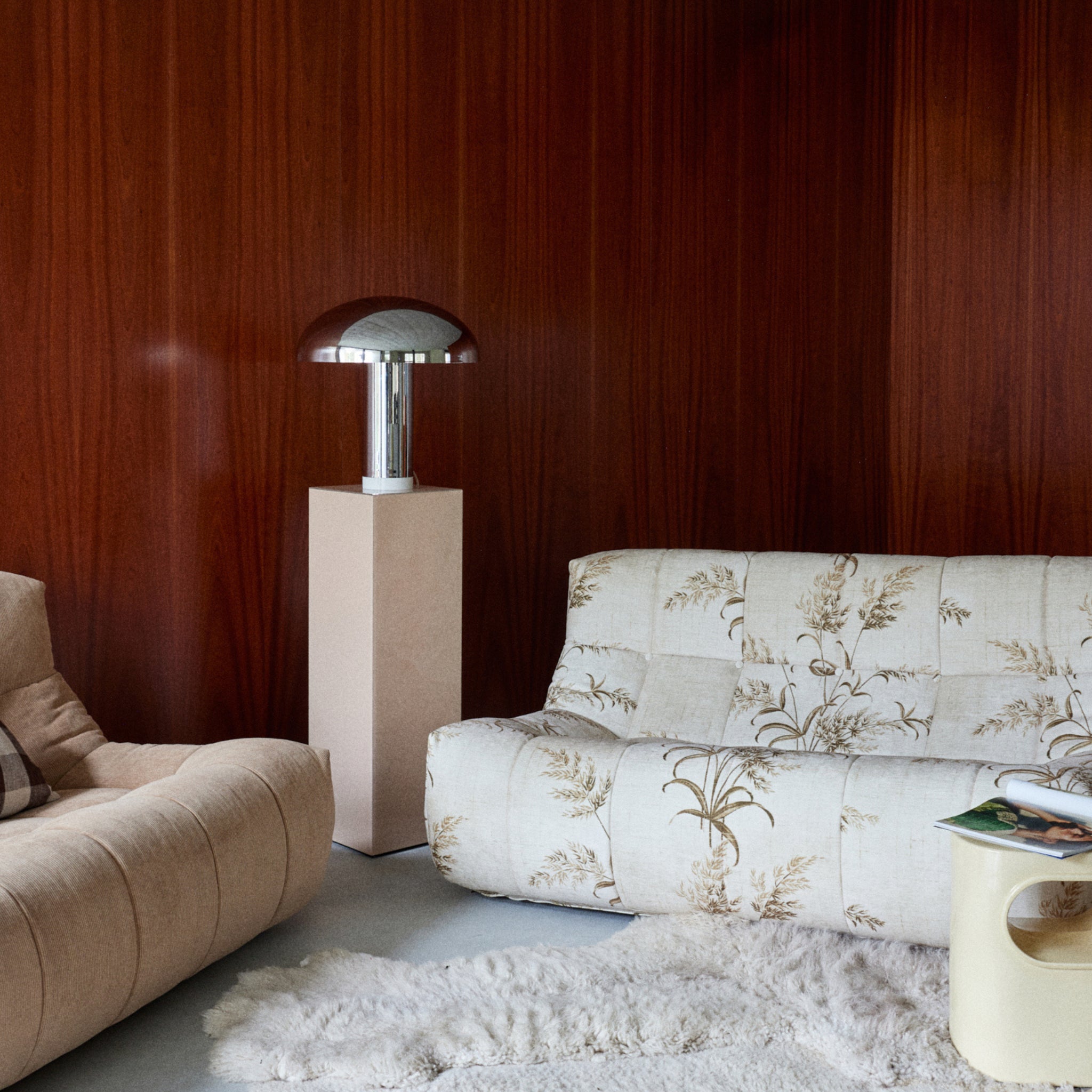 Een gezellige woonkamer met een crèmekleurige gebloemde bank, een beige fauteuil, een shaggy wit vloerkleed en de HK Living Lounge Tafellamp Chrome op een kolomtafel, allemaal tegen een rijke houten muur voor moderne warmte.