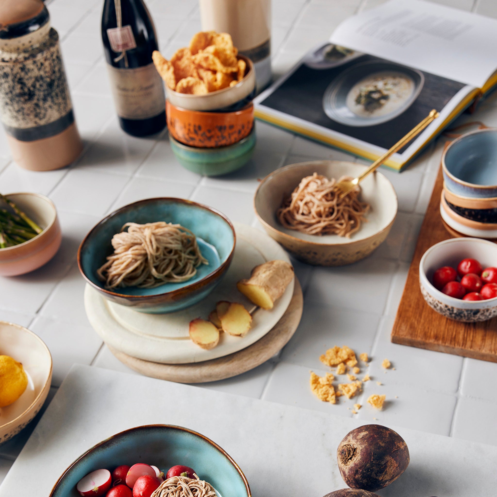 Een HK Living Serveerplank Mango van HK Living bevat keramische kommen met noedels, cherrytomaatjes, gember, chips, mogelijke mangoschijfjes, plus flessen, een kookboek en borden op een wit betegeld oppervlak.