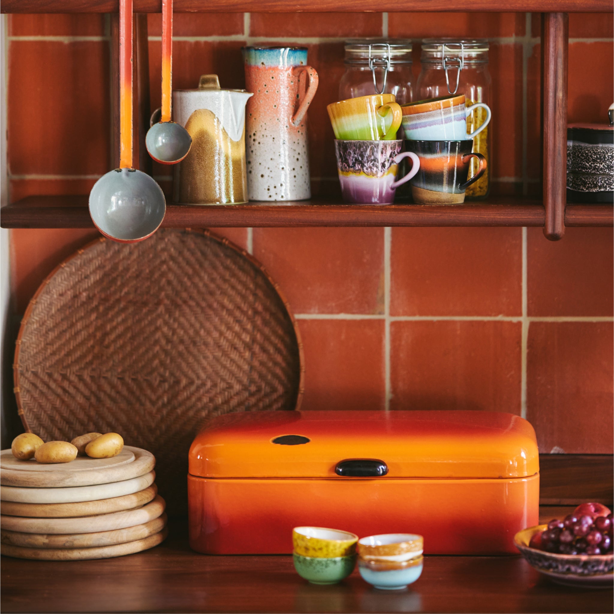Een retro keukenscène met een oranje brooddoos, gestapelde houten borden, een HK Living Serveerplank Mango serveerplank, een geweven dienblad, kleurrijke kommen, hangende maatlepels en planken met keramische mokken, potten en kruiken tegen een betegelde muur.