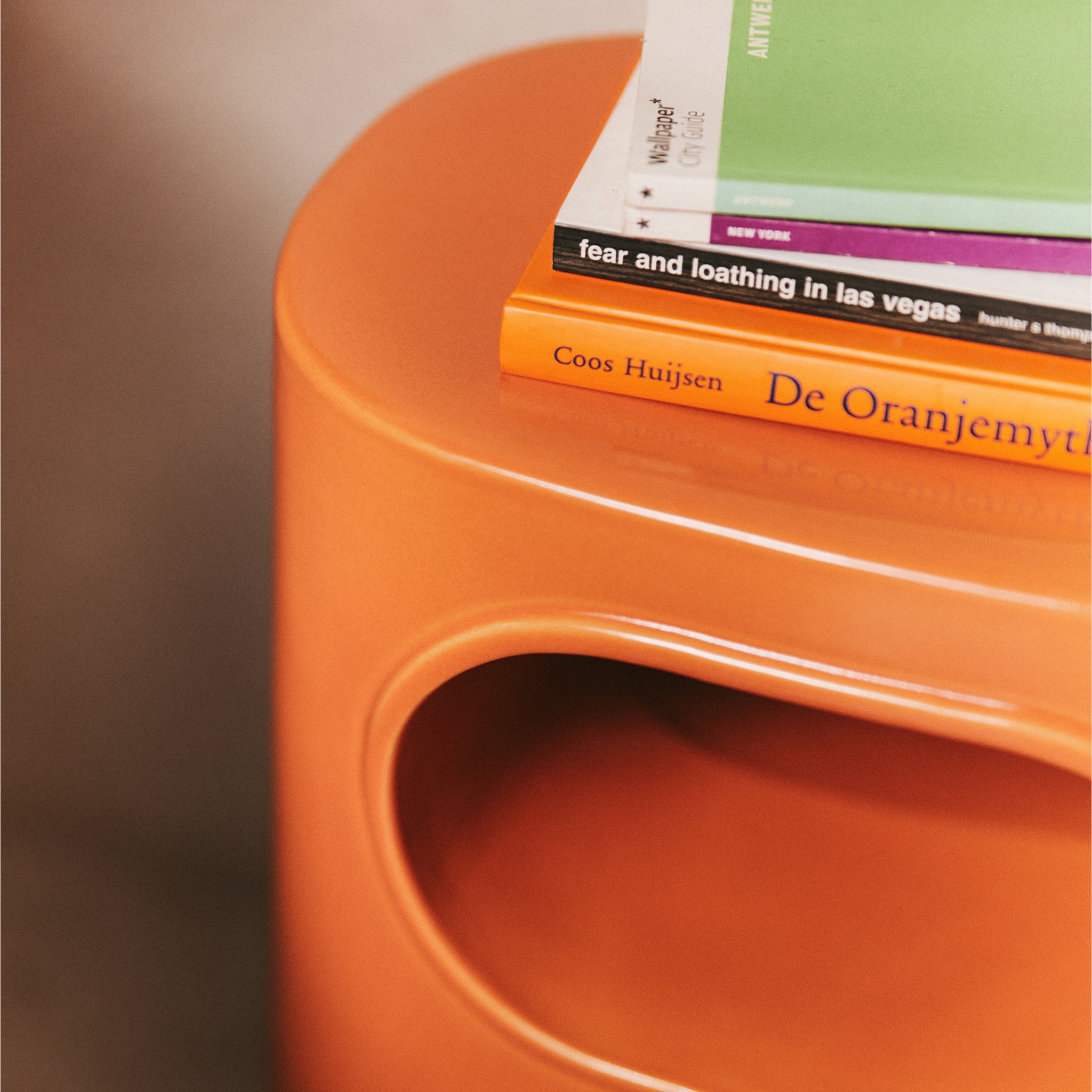 Een close-up van de HK Living Space Bijzettafel Orange van HK Living, gestyled met een stapel boeken, waaronder een met een groene kaft en het fel oranje boek De Oranjemythe van Coos Huijsen, met Space Age Design inspiratie.