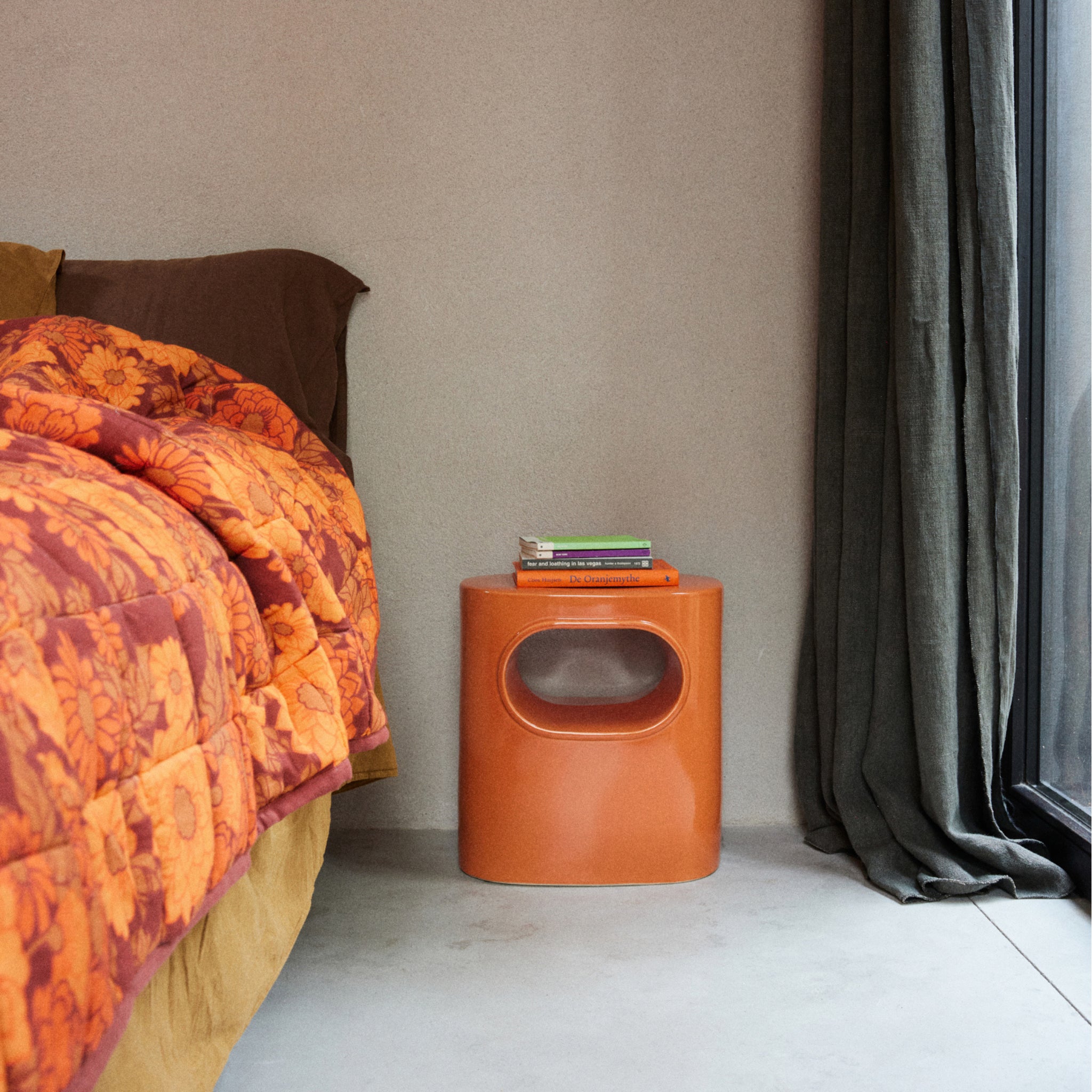 Een compacte HK Living Space Bijzettafel Orange van HK Living, met een ronde uitsparing, houdt gestapelde boeken naast een bed met een oranje gebloemd dekbed. Donkere gordijnen maken de minimalistische look met subtiele Space Age Design vibes compleet.