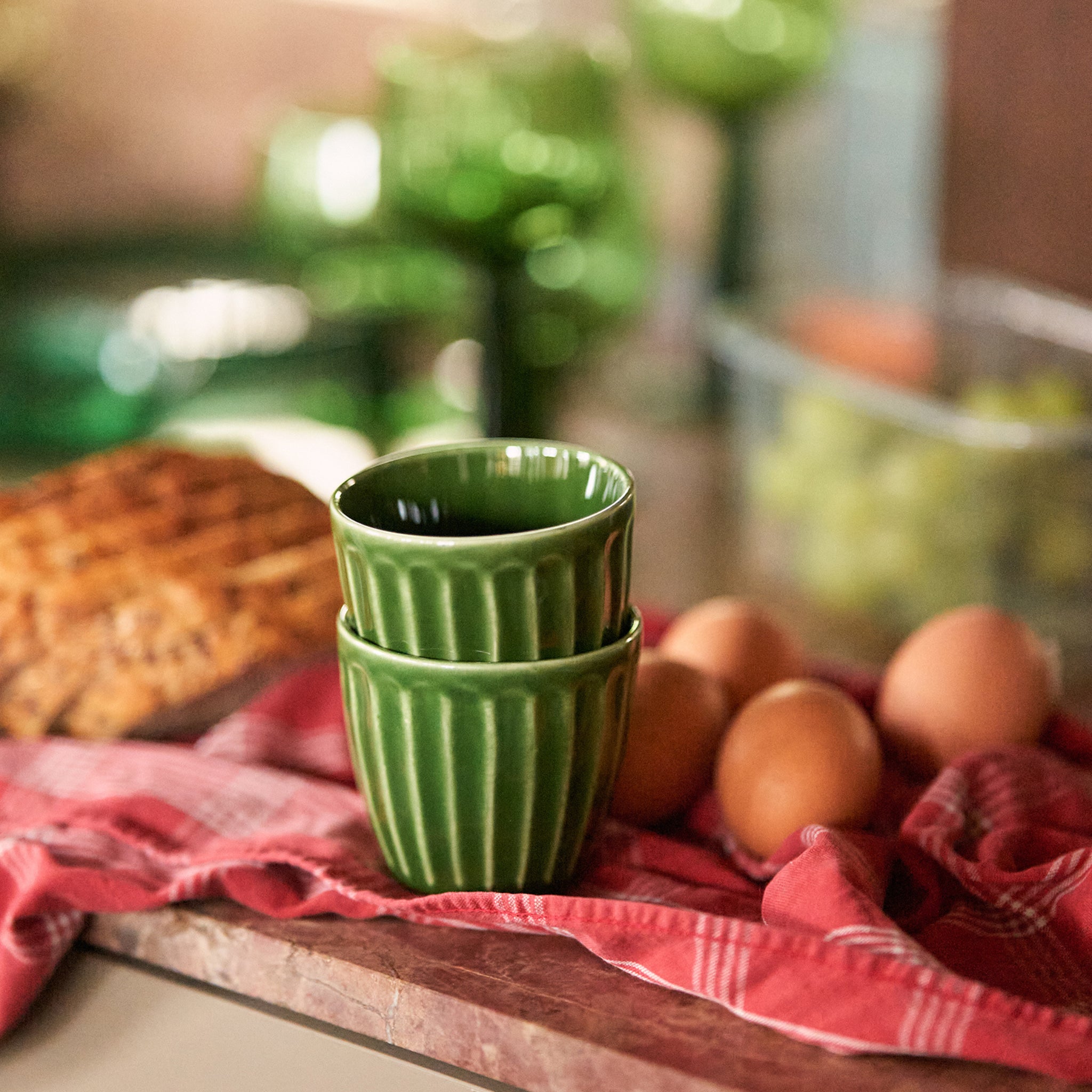 Twee HK Living The Emeralds Keramische Mok Geribbeld Groen uit een set van 4 staan op een rood geruit kleed naast eieren en brood, met vaag groen HK Living glaswerk en druiven op de achtergrond.