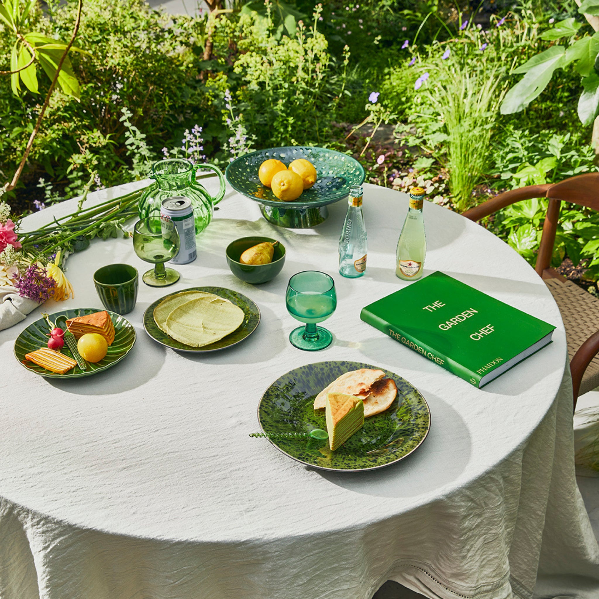 Op een ronde buitentafel met een wit kleed staan HK Living The Emeralds Wijnglas Laag Groen 4 st. glazen, borden met eten, flessen bruisend water, citroenen, bloemen en het groene boek "The Garden Chef" temidden van weelderige tuinplanten.