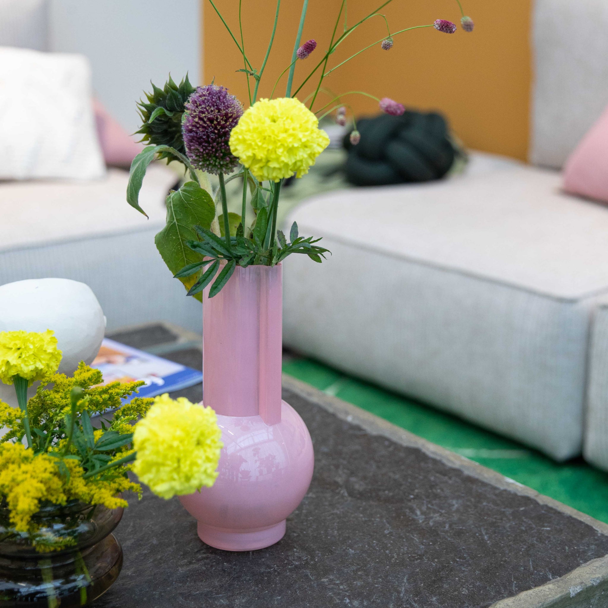 De HK Living Vaas Flamingo Roze H25 met gele en paarse bloemen siert de donkere koffietafel, naast een kleine glazen kom met gele bloemen. Beige sofa's en een groene vloer vormen de achtergrond.