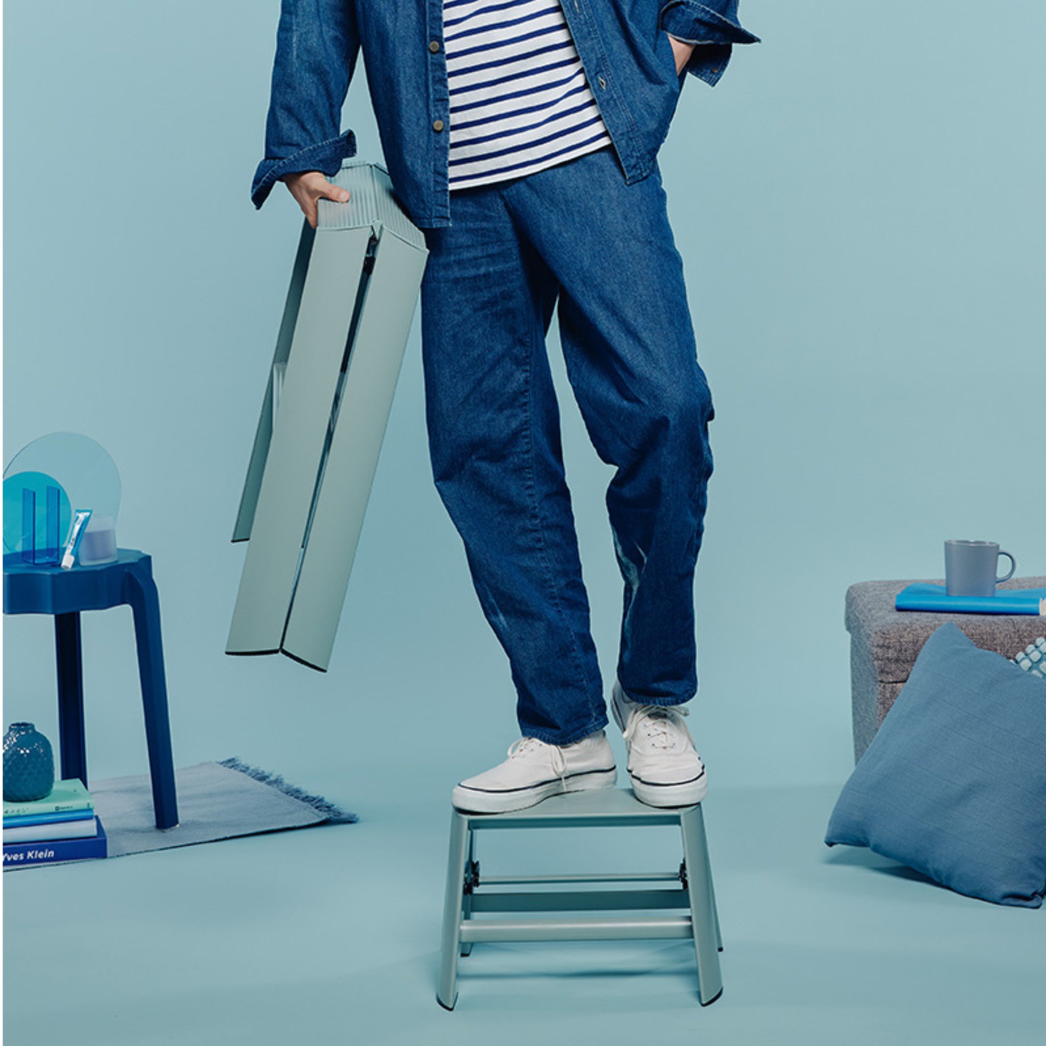 Een persoon in witte sneakers en blauwe spijkerstof staat op een Hasegawa Lucano Ladder 1 Step Dusty Mint en houdt een opgevouwen lichtblauwe tafel vast. De moderne setting heeft rustgevende blauwe tinten en strakke, minimale designelementen van Hasegawa.