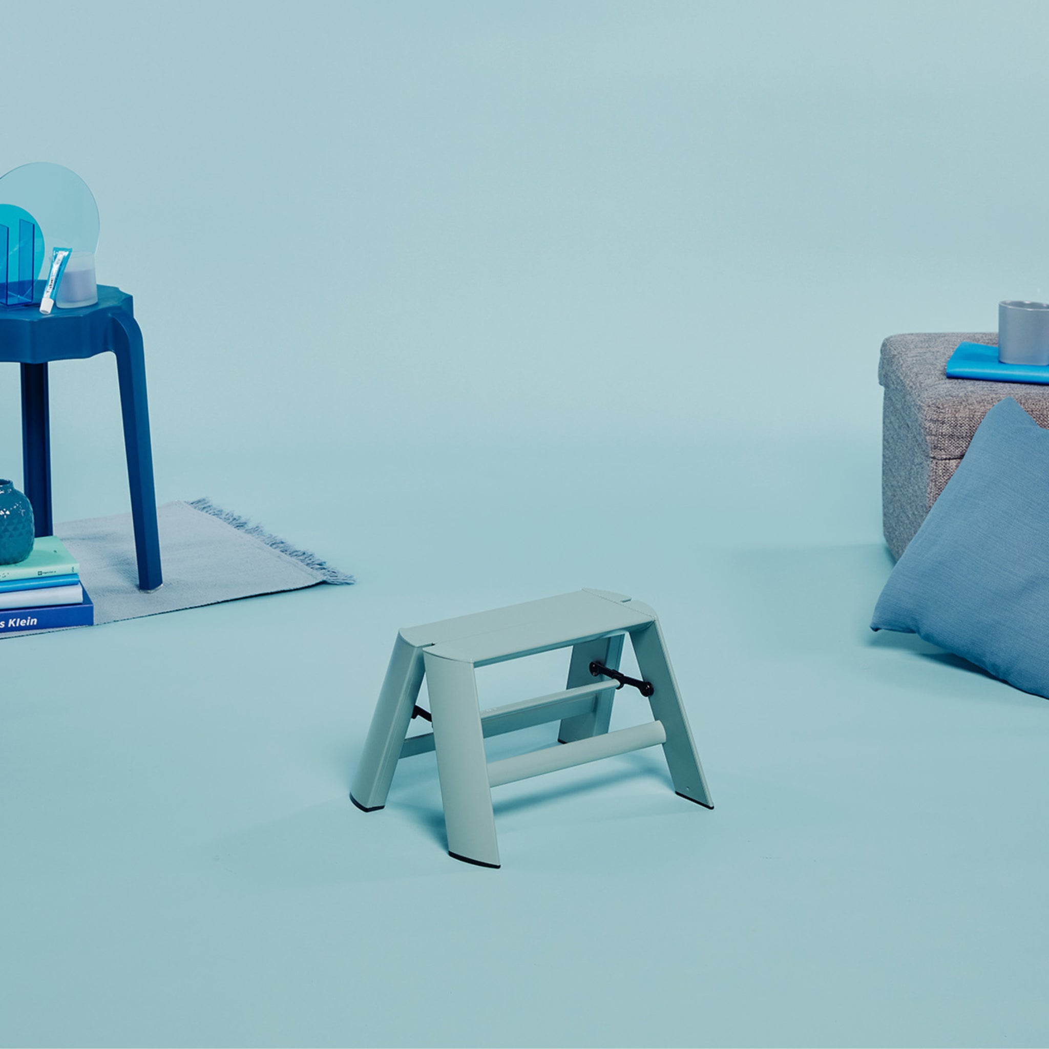 Een Hasegawa Lucano Ladder 1 Trede in Dusty Mint voegt een stijlvolle touch toe aan het blauw opstapje, gecombineerd met een blauwe tafel, boeken, decor, grijs voetenbankje en blauw kussen voor een samenhangende look.