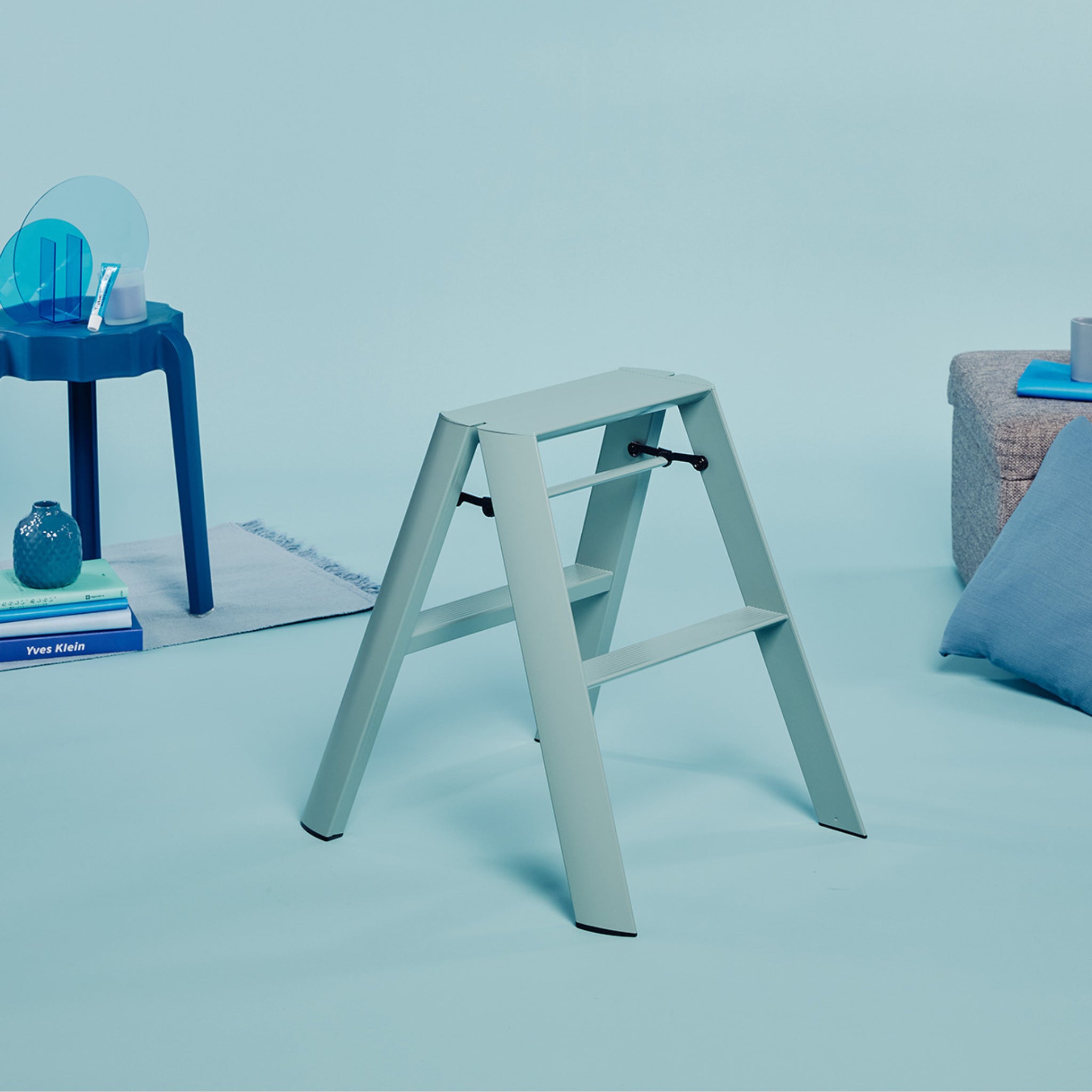Een Hasegawa Lucano Ladder 2 Step in stoffig mint van Hasegawa staat op een lichtblauwe vloer, vergezeld door een blauwe bijzettafel met boeken en decor, een grijze zitkussens en een lichtblauw kussen in een minimalistische Japans geïnspireerde omgeving.