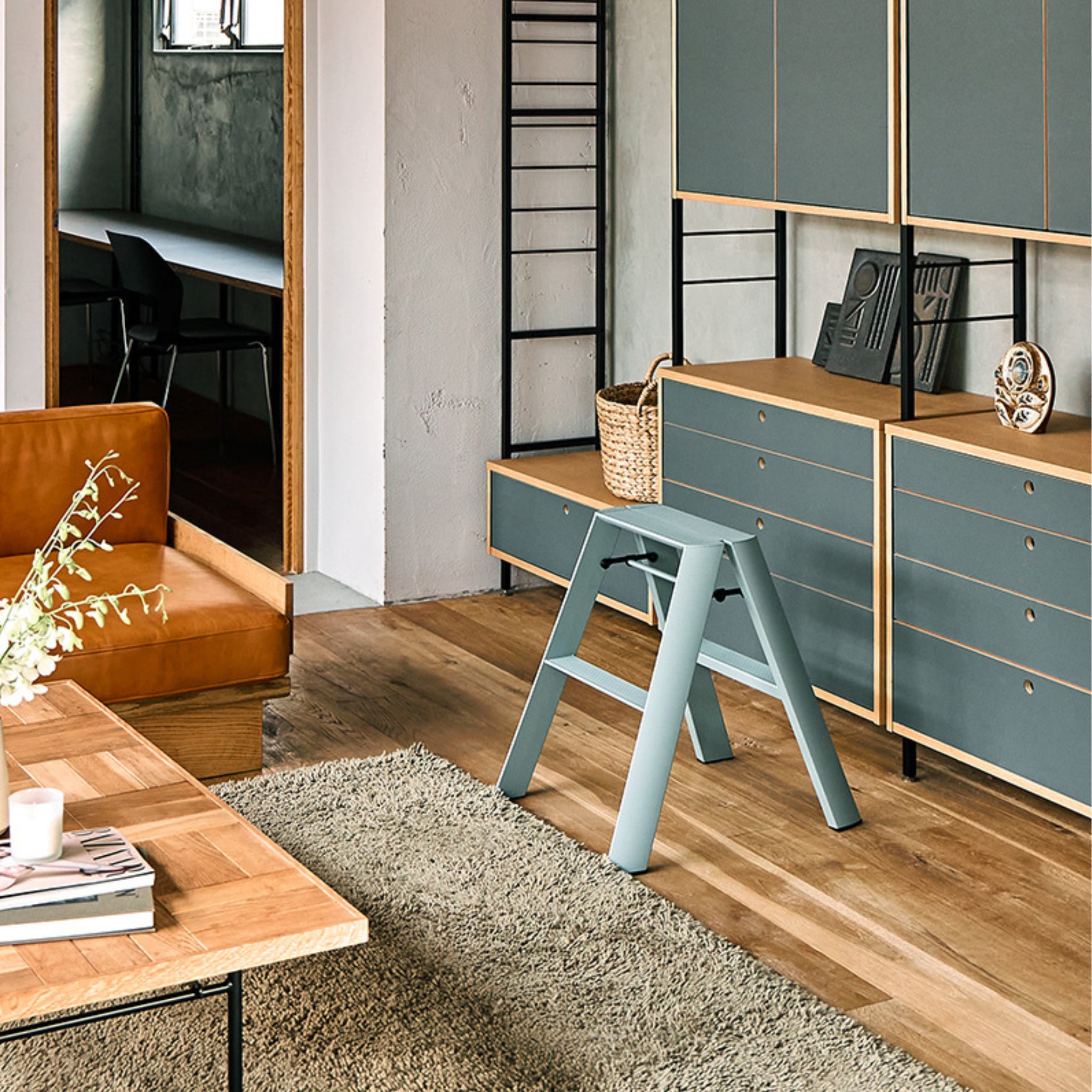 Moderne leefruimte met houten en blauwe kasten, een bruine leren bank, lichthouten salontafel en beige vloerkleed. De Hasegawa Lucano Ladder 2 Step in Dusty Mint staat in het middelpunt van de belangstelling en benadrukt het minimale Japanse ontwerp. Deuropening onthult bureau en stoel.