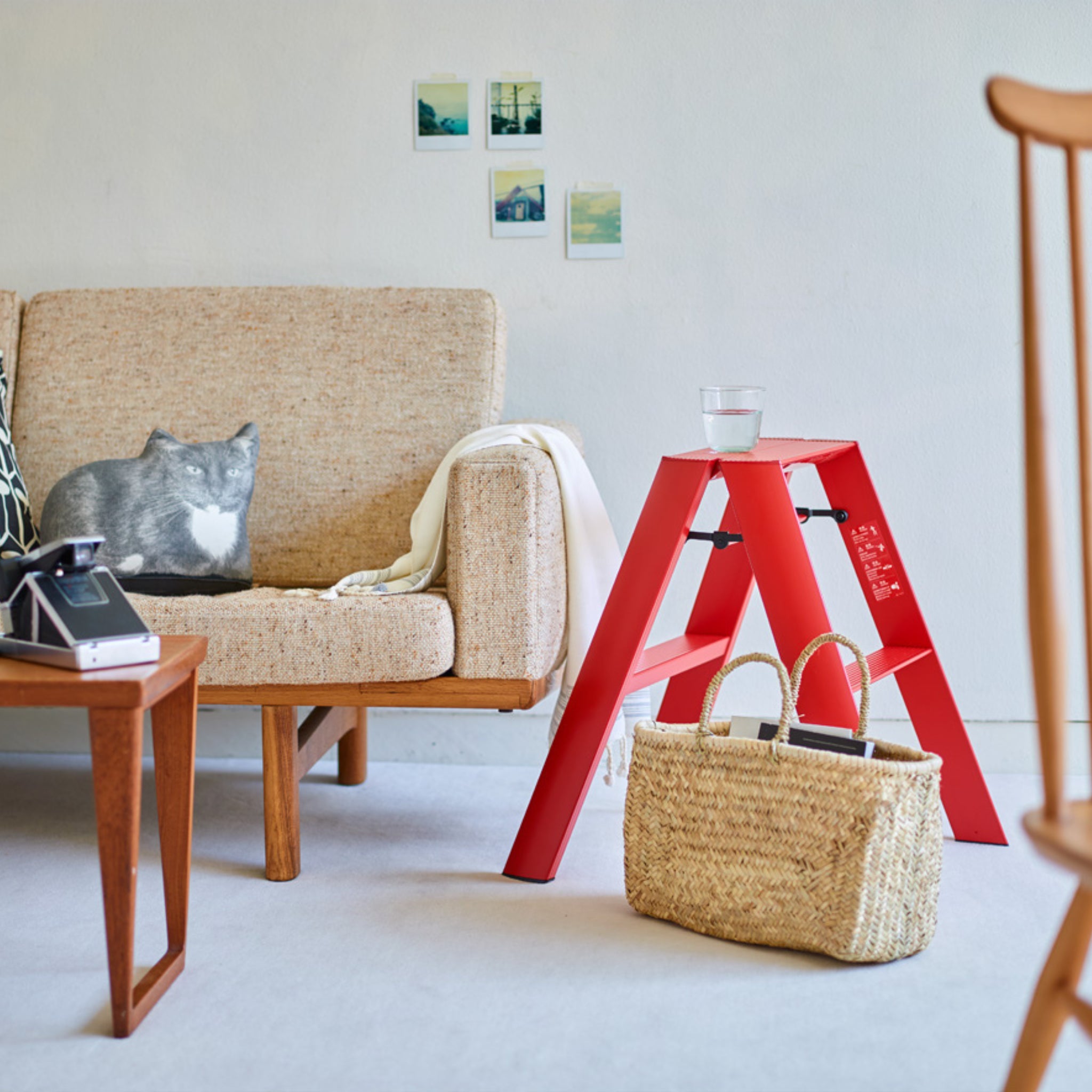 Een gezellige woonkamer met een beige bank, een kattenkussen, een deken, een houten tafel met camera en een rode Hasegawa Lucano Ladder 2 Step Rood die een glas water vasthoudt, plus een geweven rieten zak en kleine schilderijtjes op de witte muur.