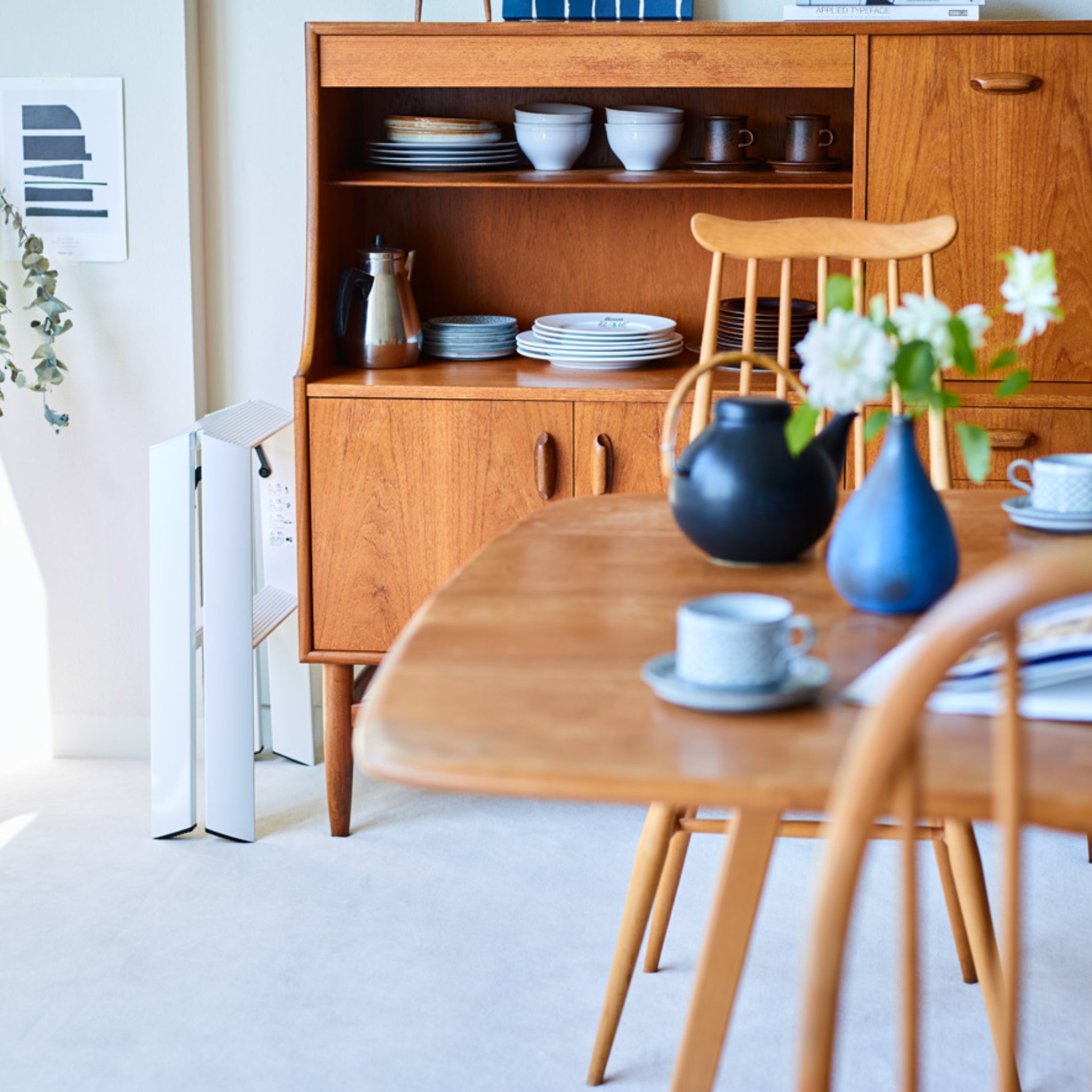 Een lichte eethoek met houten meubels toont Japanse invloeden, geaccentueerd door een zwarte theepot en blauwe vaas. De Hasegawa Lucano Ladder 2 Step Wit staat vlakbij, terwijl kunst en planten de uitnodigende sfeer compleet maken.