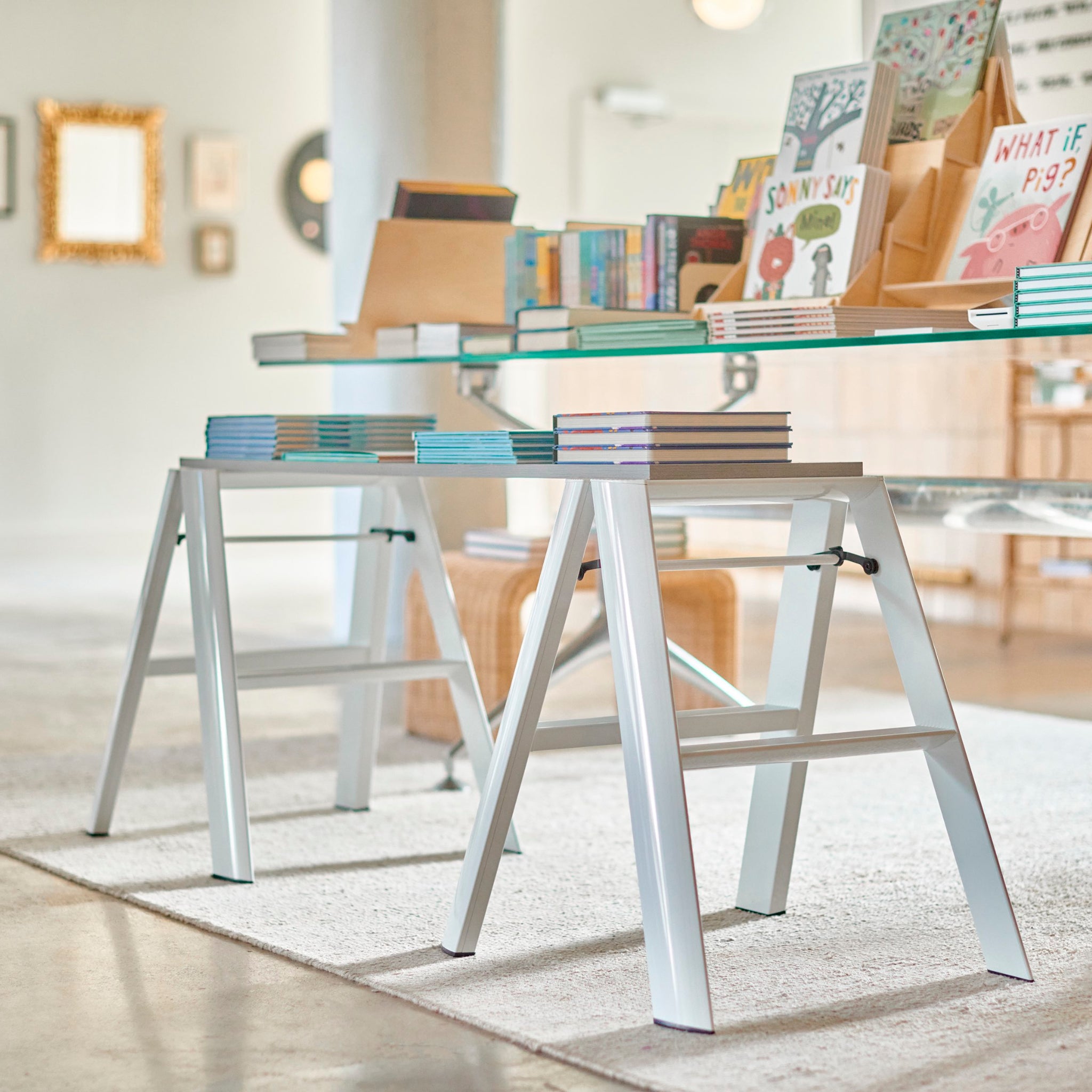 Een moderne boekwinkel toont levendige stapels boeken op een glazen tafel met witte zaagbokpoten in een lichte kamer versierd met kunst, planken en de strakke Hasegawa Lucano Ladder 2 Step Wit van Hasegawa geïnspireerd op Japans design.