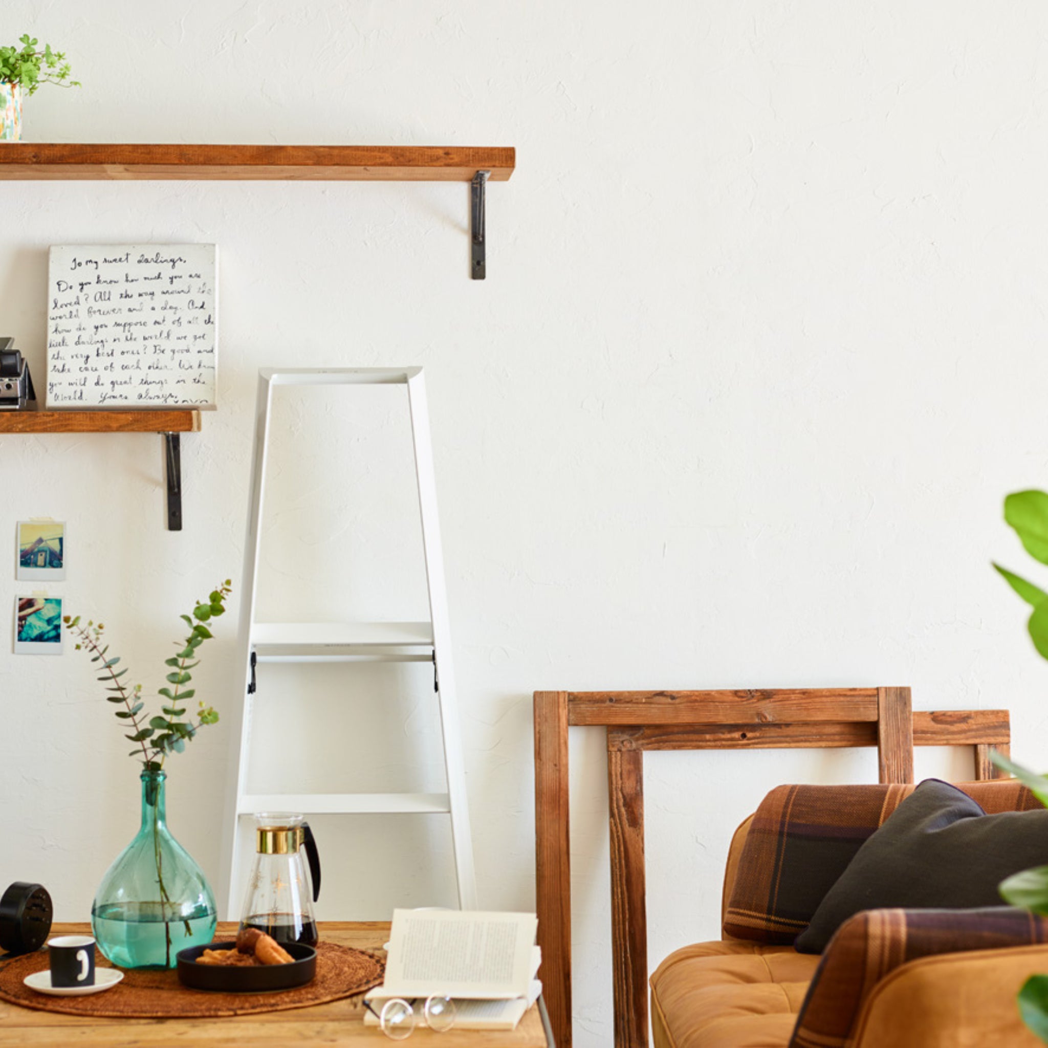 Een gezellige woonkamer heeft een bruine bank, houten planken aan een witte muur, ingelijste kunst en subtiel Japans design. De Hasegawa Lucano Ladder 3 Step Wit van Hasegawa staat dichtbij, terwijl drankjes en boeken op het ronde dienblad van de houten tafel staan.