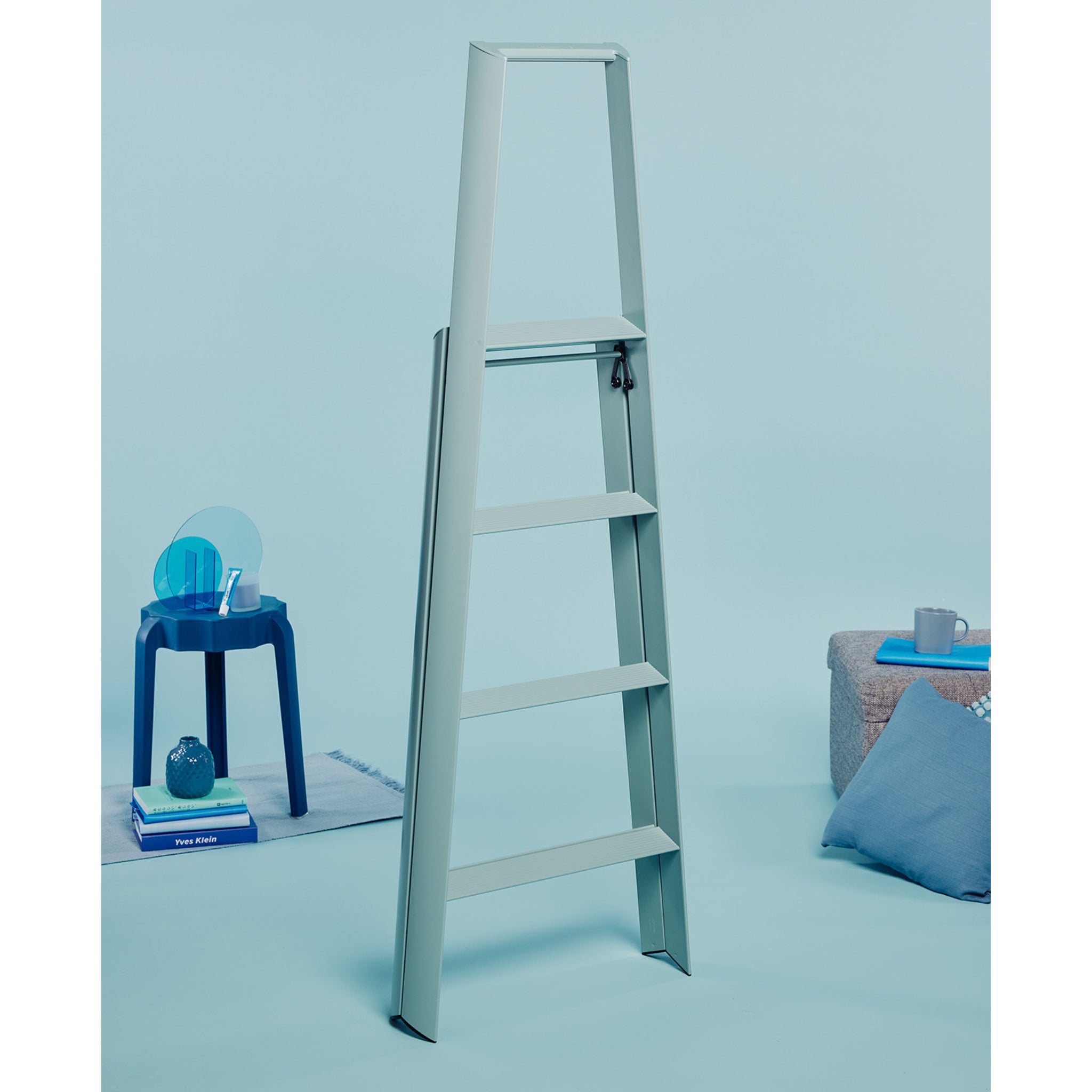 De Hasegawa Lucano Ladder 4 Stap Dusty Mint van Hasegawa staat rechtop in een minimalistische blauwe kamer. Een kleine blauwe tafel met boeken en decor, plus een grijs kussen met een kopje, creëren een rustige omgeving.