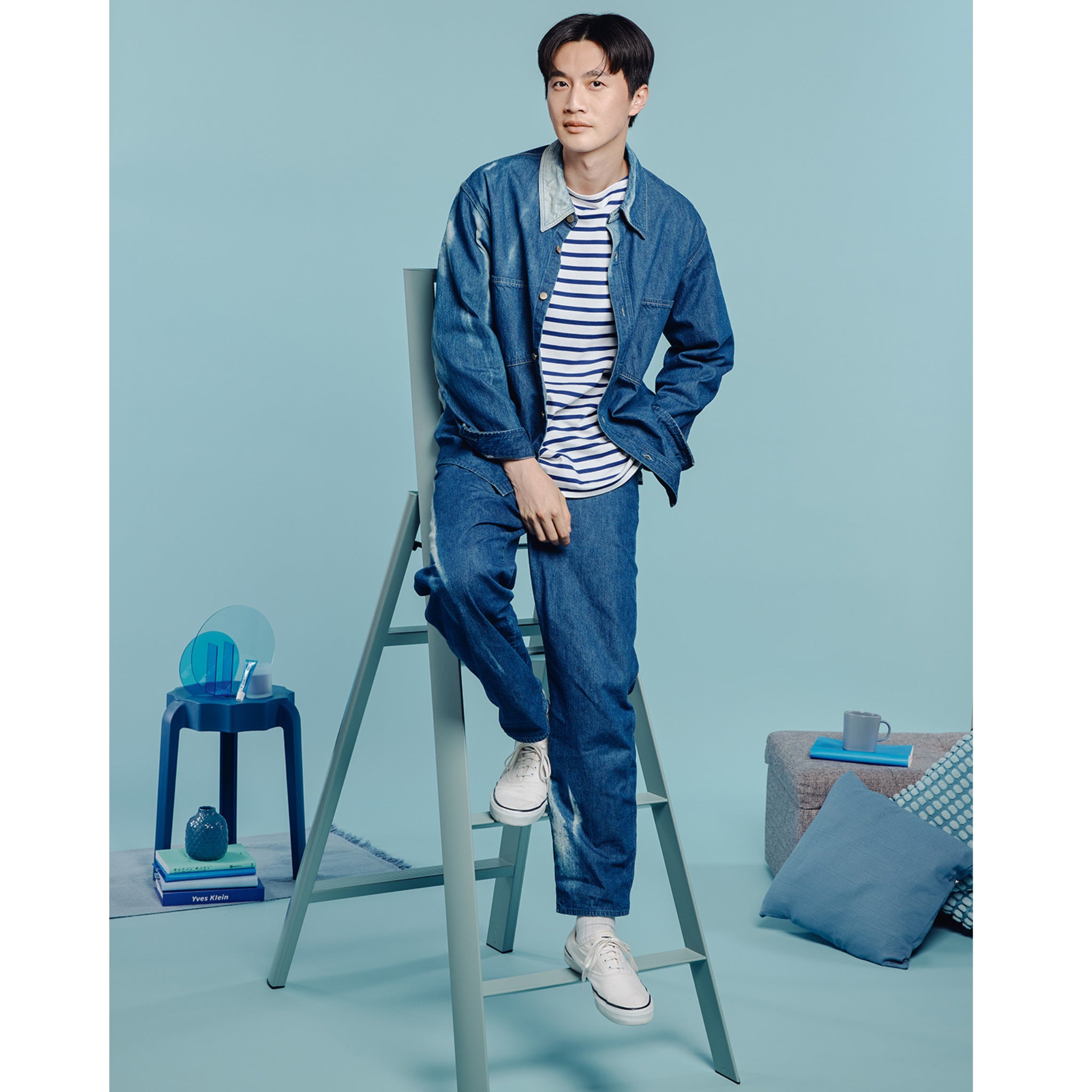 Een jonge man in een spijkerjack, spijkerbroek, gestreept shirt en witte sneakers zit op een Hasegawa Lucano Ladder 4 Step Dusty Mint van Hasegawa tegen een blauwe achtergrond, omringd door blauw decor zoals een tafel en kussens.