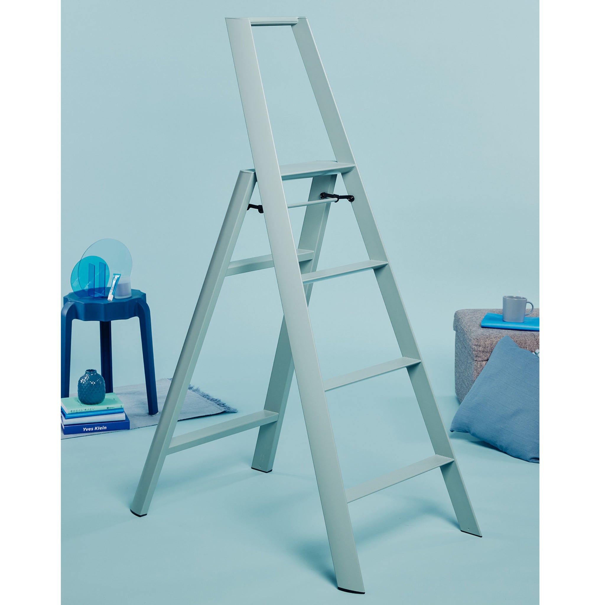 Een Hasegawa Lucano Ladder 4 Step in Dusty Mint van Hasegawa staat in een minimalistische kamer met blauwe muren, een bijpassende kruk met een lamp en boeken en een grijze poef met daarop een mok, boek en blauw kussen.