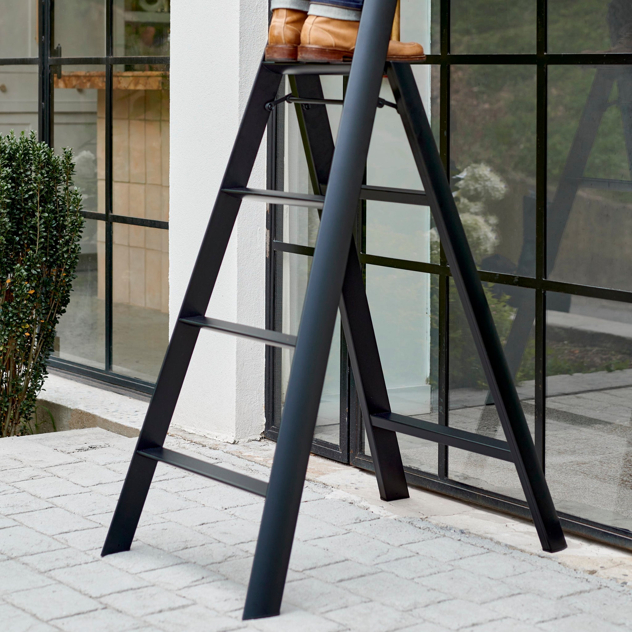 Een persoon staat op een zwarte Hasegawa Lucano Ladder 4 Step Rood buiten een gebouw met grote glazen ramen en een betegeld stenen terras, waarbij alleen het onderste gedeelte van de ladder en hun benen en laarzen zichtbaar zijn.