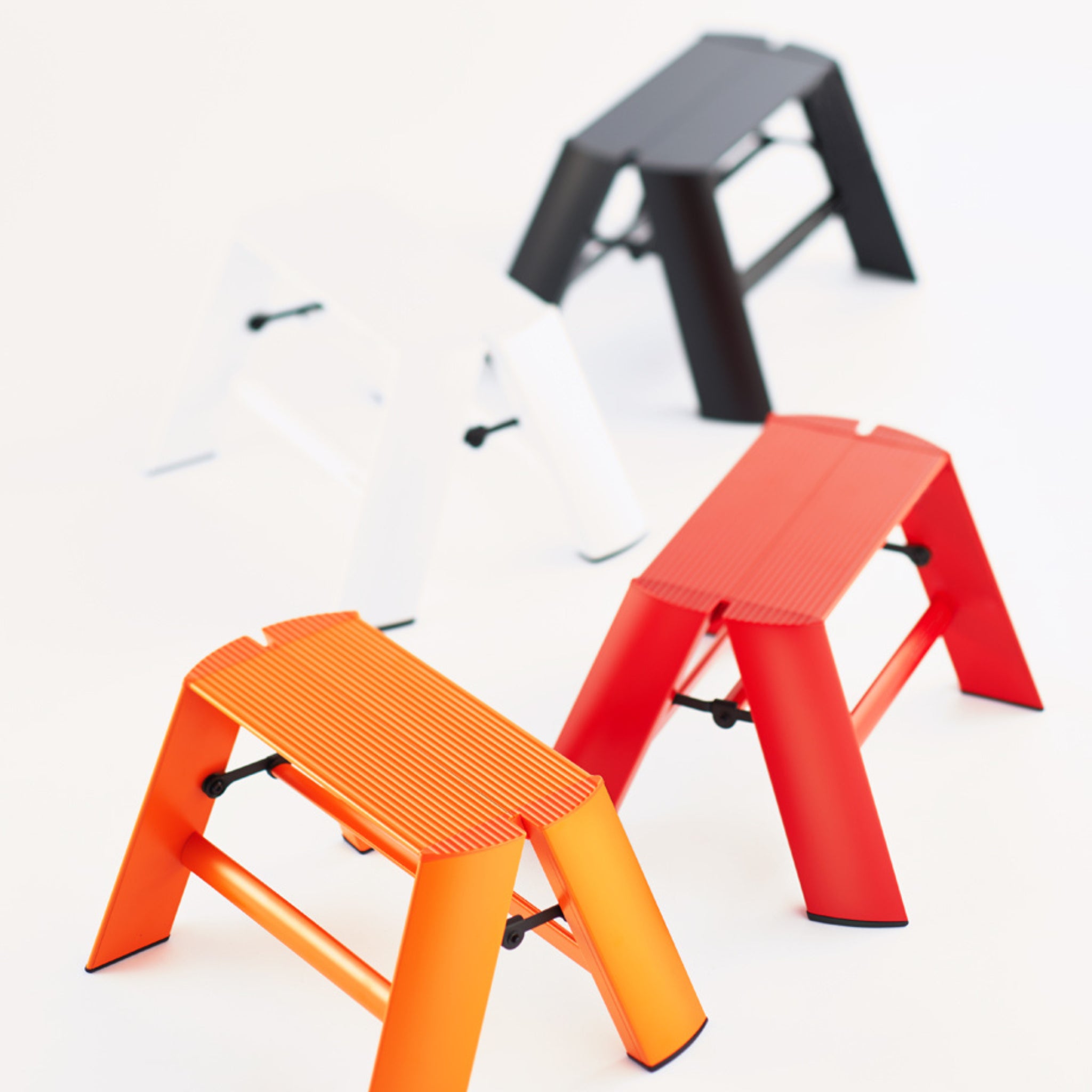 Vier compacte Hasegawa Lucano Ladder 1 Step krukjes in oranje, rood, wit en zwart staan op een witte achtergrond. Elk minimalistisch opstapje van Hasegawa heeft schuine poten en geribbelde zittingen voor stijl en stabiliteit.