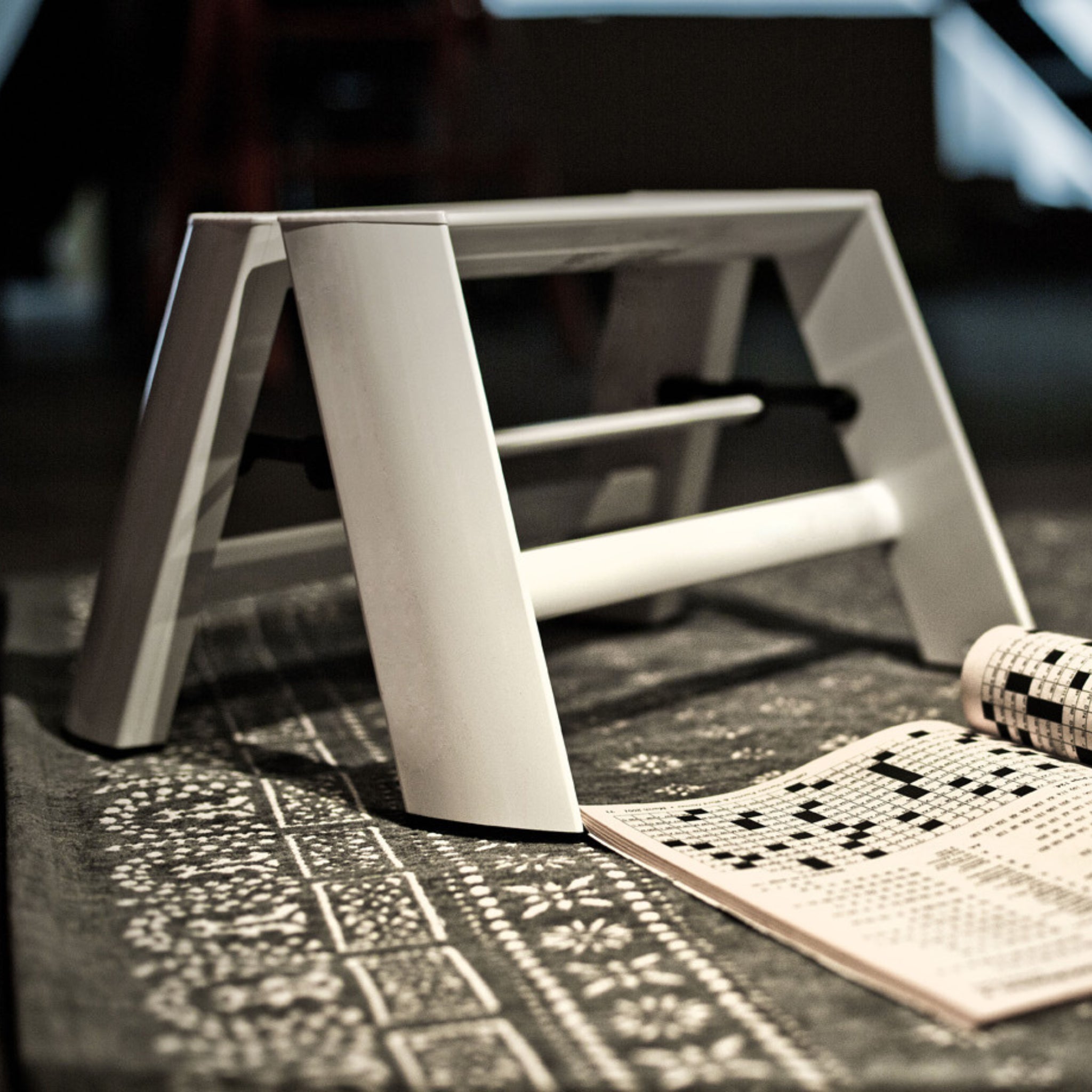 Een Hasegawa Lucano Ladder 1 Stap Wit van Hasegawa, geïnspireerd op Japans design, staat op een vloerkleed met patroon naast een open boek met kruiswoordraadsels, waarbij zachte verlichting een gezellige sfeer creëert.