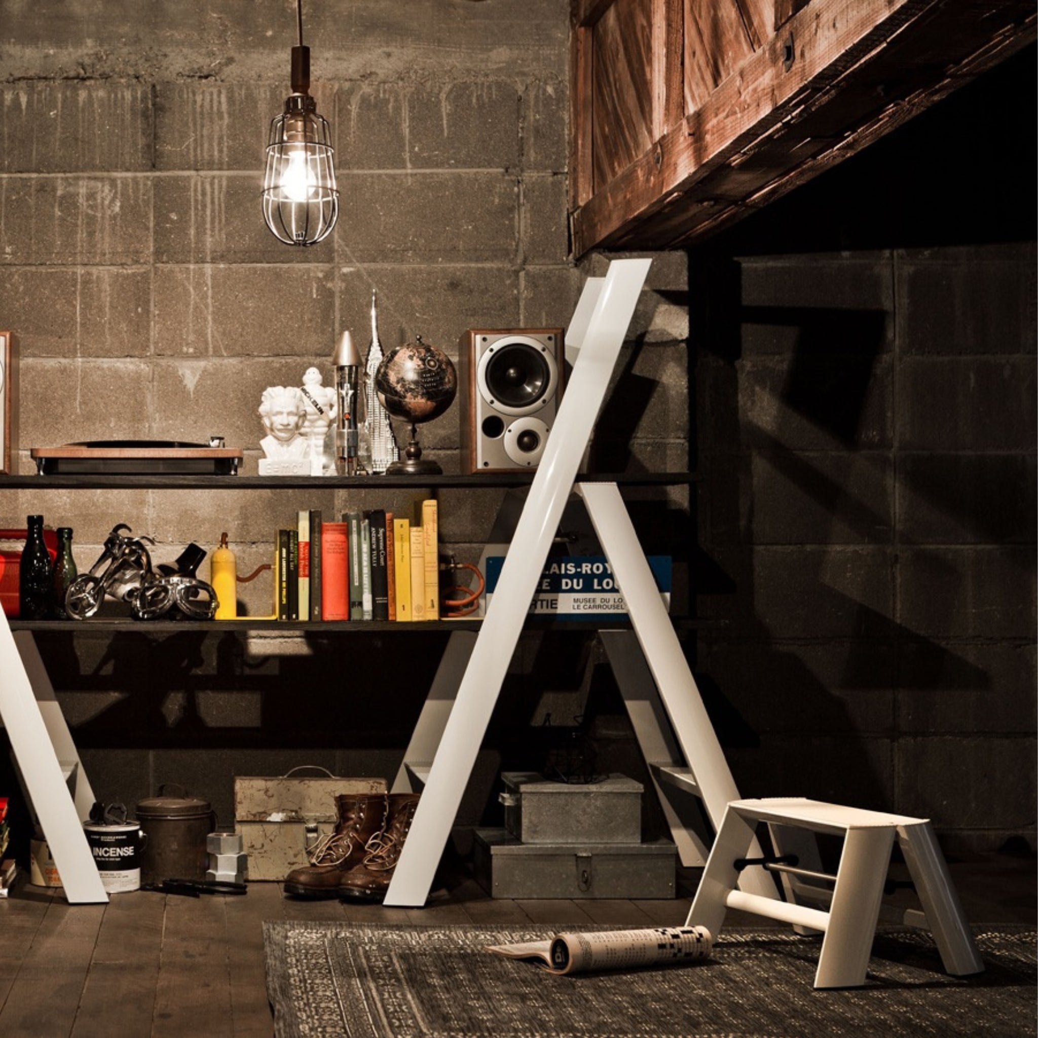 Een gezellige werkkamer in industriële stijl met een houten bureau, boeken, decor, luidspreker, modelraket, laarzen, opbergdozen en een Hasegawa Lucano Ladder 1 Step Wit van Hasegawa ligt op een vloerkleed onder warme hanglampen.