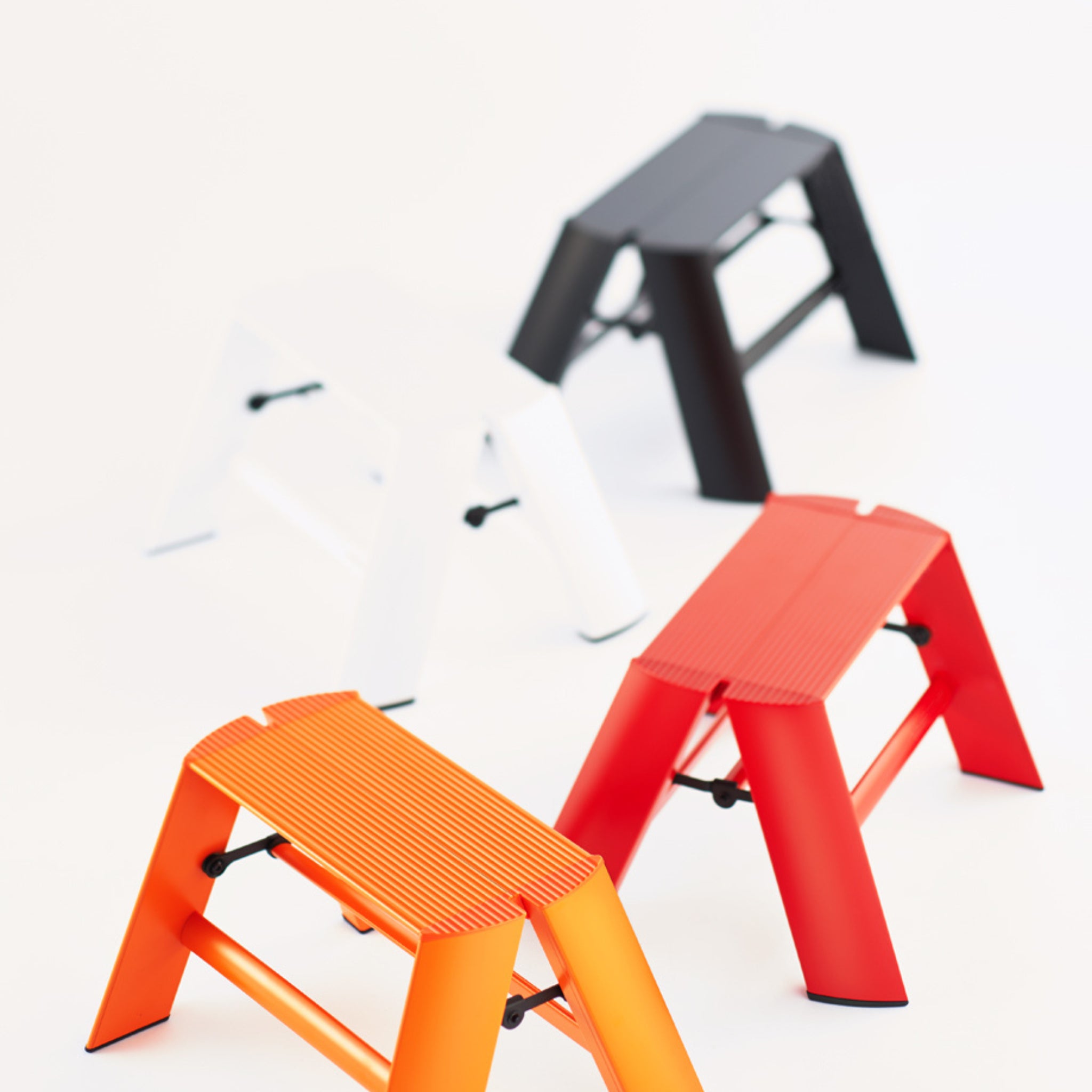 Vier Hasegawa Lucano Ladder 1 Step krukjes - oranje, rood, wit en zwart - met Japans design staan diagonaal opgesteld op een wit oppervlak, met het oranje krukje vooraan en het zwarte (Zwart) krukje achteraan.