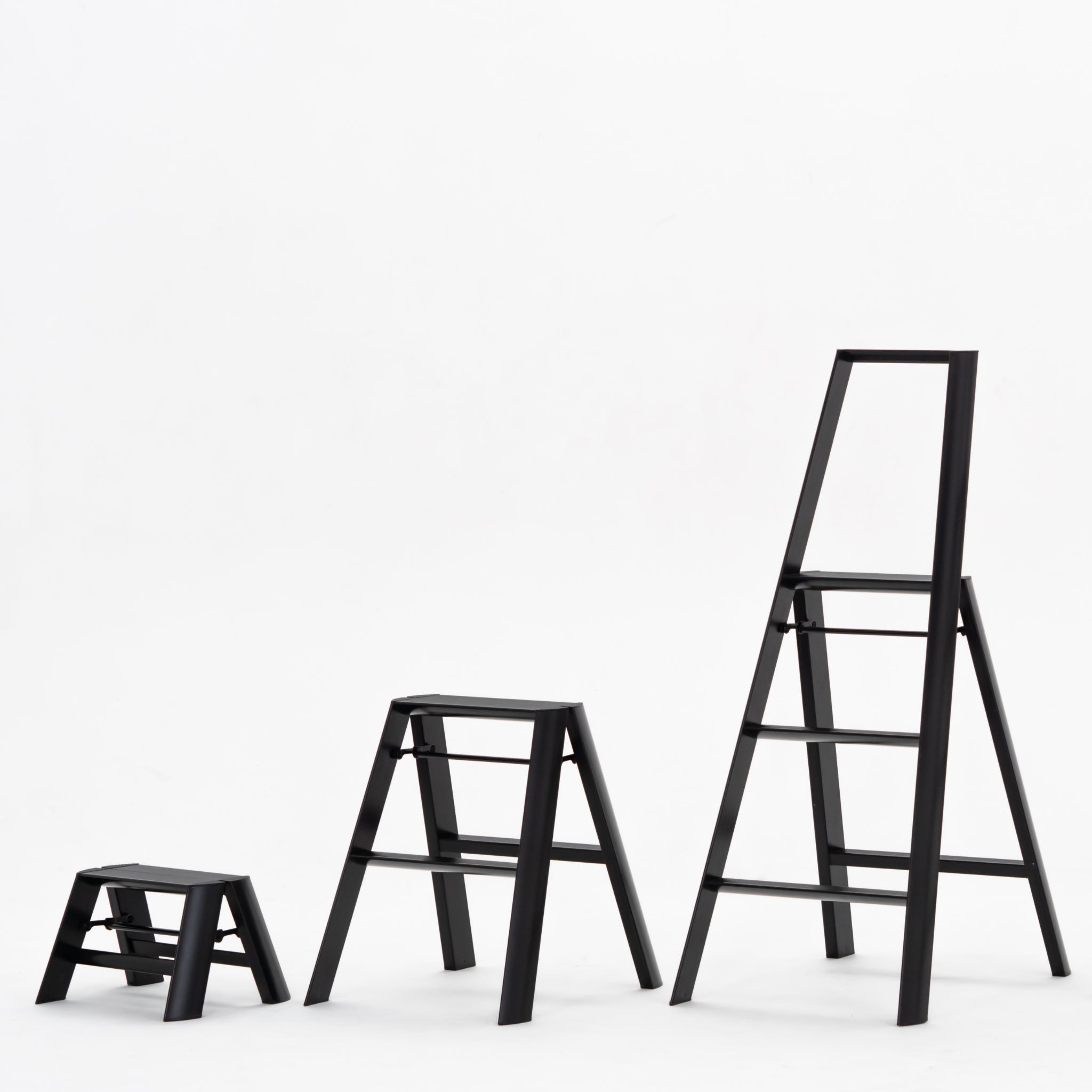 Drie zwarte Hasegawa Lucano Ladder 1 Stap Zwart ladders van Hasegawa staan in een rij op een effen witte achtergrond, waardoor hun compacte Japans geïnspireerde ontwerp en variërende hoogtes van de kortste naar de hoogste benadrukt worden.
