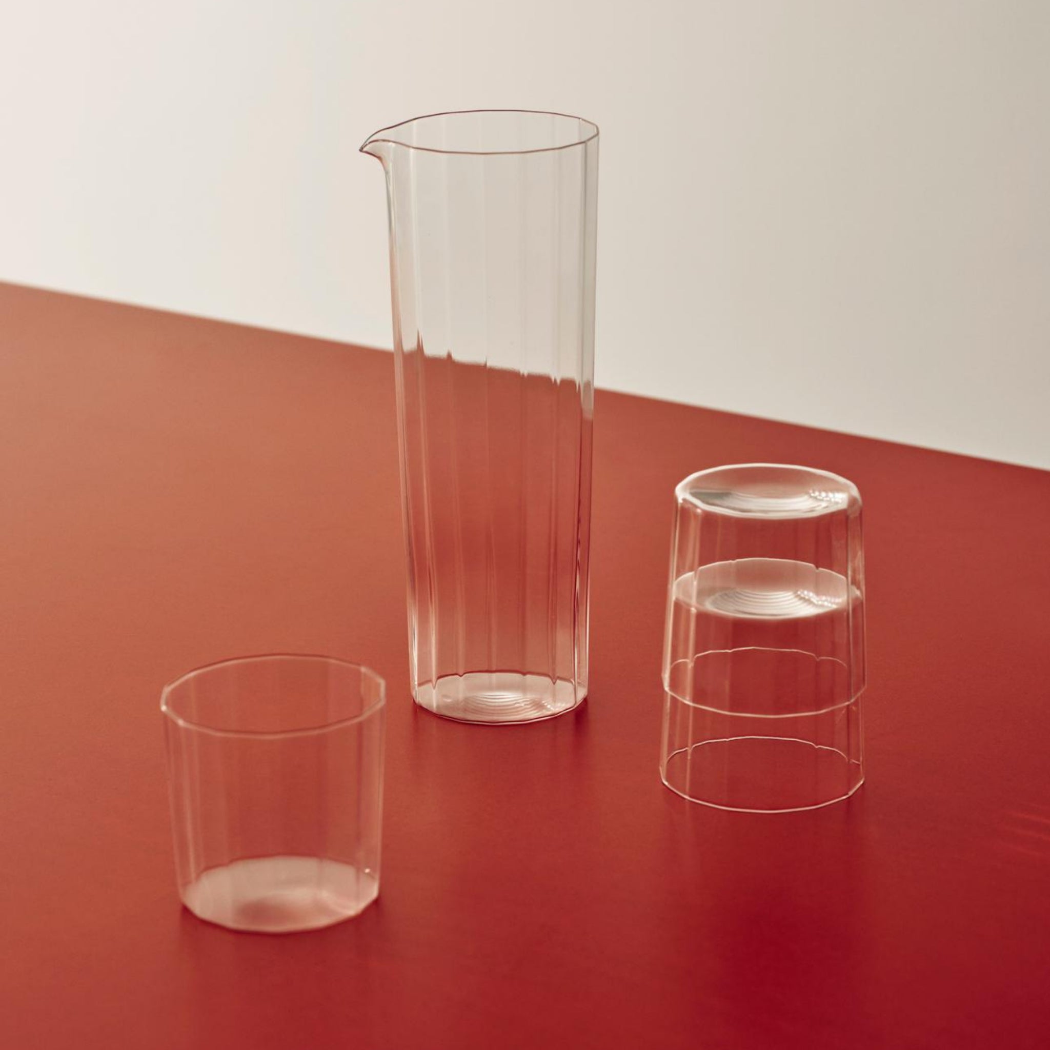 Drie heldere, geribbelde Hay Angle Waterglazen 4 st. van Hay staan op een rood oppervlak - één rechtop, één ondersteboven en één hoge kan - gemaakt van kristallijn glas voor een elegante look tegen een gebroken witte achtergrond.