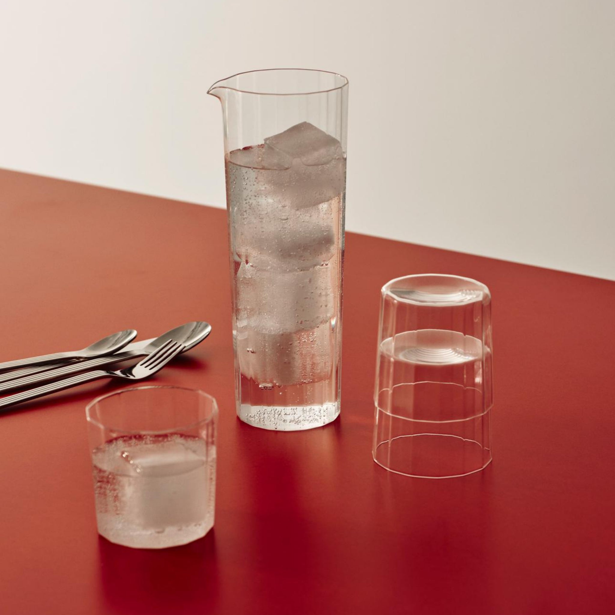 Een set Hay Angle Waterglazen (4 stuks) van Hay, een gevuld met ijswater en een andere met een ijsblokje, naast een omgekeerd leeg glas en bestek, rusten op een rood tafelblad voor een elegante look tegen een witte achtergrond.
